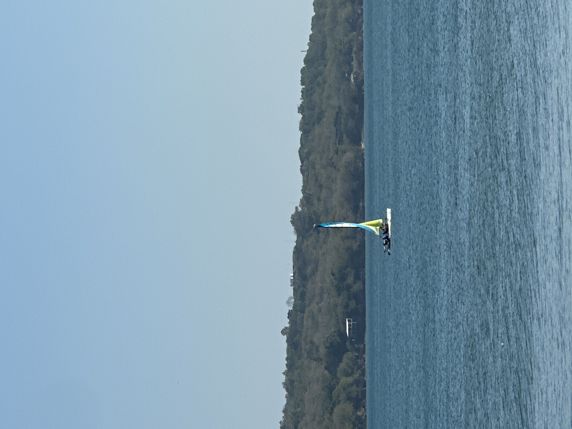 A sailboat with a blue, yellow, and white sail is on the water. The boat has two people on it. In the background, there is a hill covered in trees and some buildings. The sky is clear and blue.
