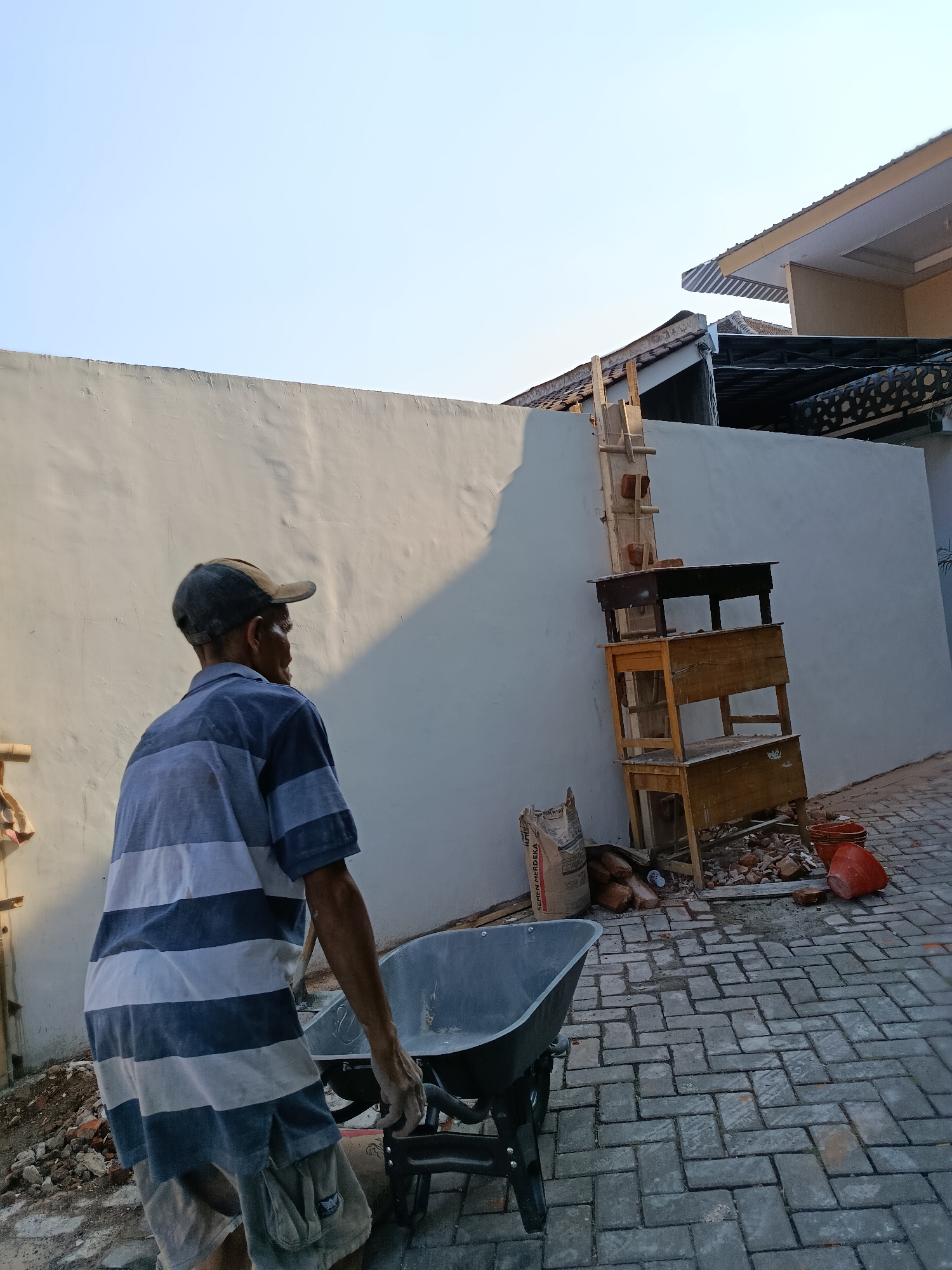 An elderly man wearing a blue and white striped shirt and a baseball cap pushes a wheelbarrow on a paved walkway. The walkway is made of grey interlocking bricks. To the left of the man, a white wall casts a long shadow. To the right of the wall, a wooden ladder is propped up against the wall, with several wooden structures stacked underneath it. A bag of cement is on the ground near the wheelbarrow, and a few orange buckets are visible near the wooden structures. The sky is clear and blue. The scene appears to be outdoors in a residential area. The specific location within Bondowoso, Indonesia, cannot be determined from the image alone, but the style of the house and the surrounding environment suggest a warm, tropical climate.