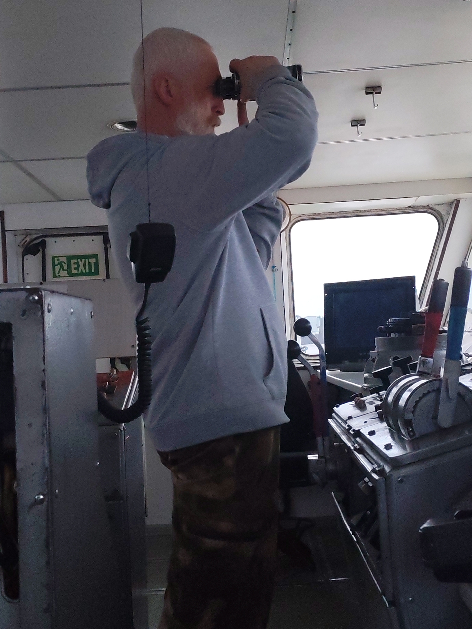 The image depicts a man on the bridge or in the control room of a marine vessel, likely in Ankhimovo, Russia. The main subject is a middle-aged man, seen from the side, facing right. He has a bald head and a prominent white beard. He is wearing a light grey hooded sweatshirt and dark, possibly patterned, trousers.
The man is actively engaged in looking through a pair of black binoculars, holding them up to his eyes with both hands. His gaze is directed out of a large, rectangular window, which dominates the right side of the frame and reveals a bright, uniformly lit exterior, suggesting an overcast daytime sky.
To the left of the man, a black microphone with a coiled cord hangs down, indicating communication equipment. Behind him, a green "EXIT" sign with a white running man pictogram and white text is clearly visible on a metal wall. The interior setting features utilitarian grey metallic panels forming the walls and structural elements. The ceiling is white, paneled, and has two small hooks hanging down.
In the foreground, to the man's right, is a control console or dashboard. This console features several levers, some with red and blue caps, as well as various other metallic controls and a partially visible dark monitor screen facing away from the viewer. The scene suggests a focused professional on duty aboard a ship, possibly observing for navigation or reconnaissance in a waterway context relevant to Ankhimovo.