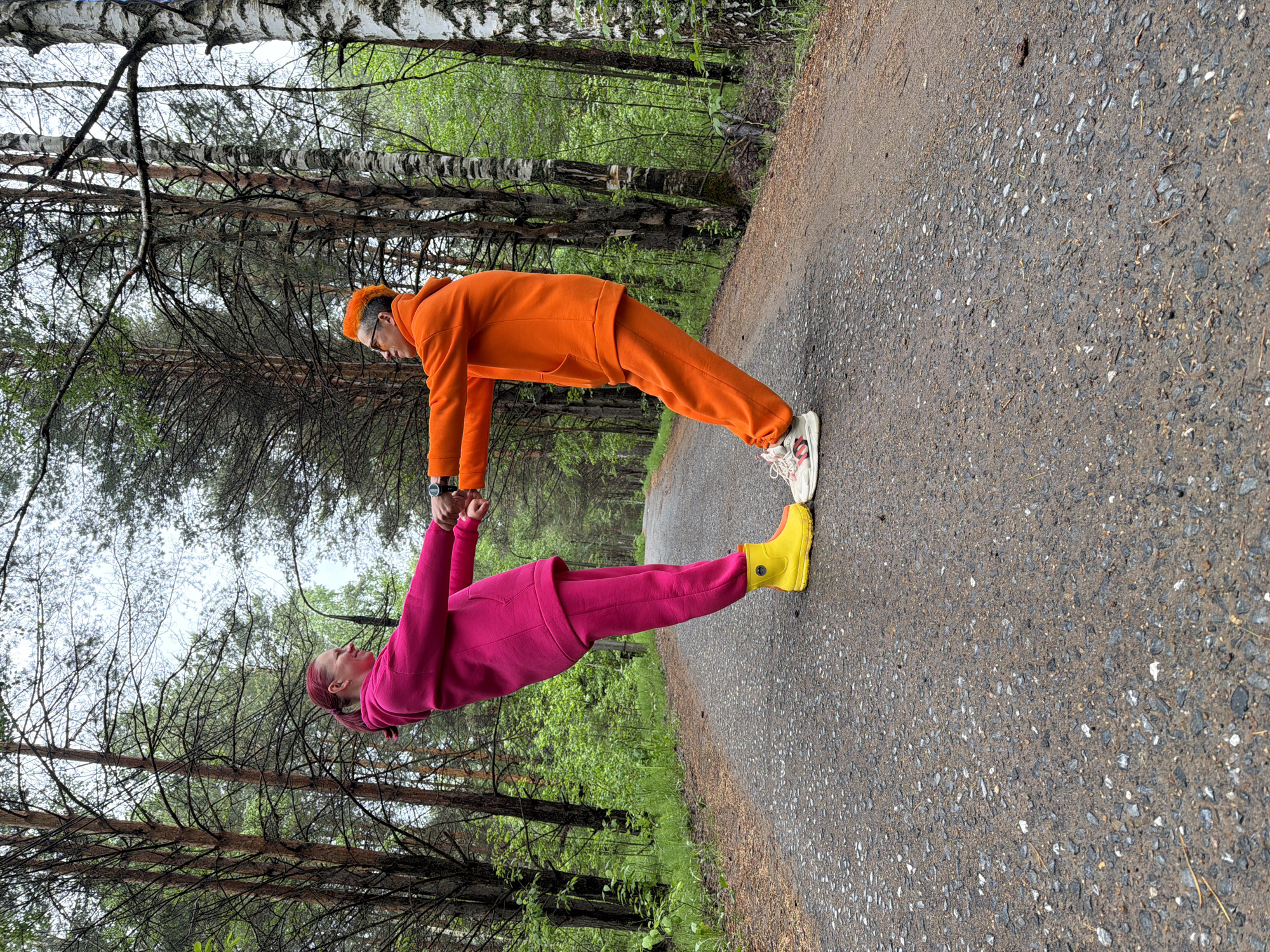 Two people, one in a pink tracksuit and yellow boots, the other in an orange tracksuit and white sneakers, form a right angle with their bodies on a paved path. They are holding hands at the vertex of the angle. A forest is visible in the background. The image was taken in the city of Chaikovsky, Russia.