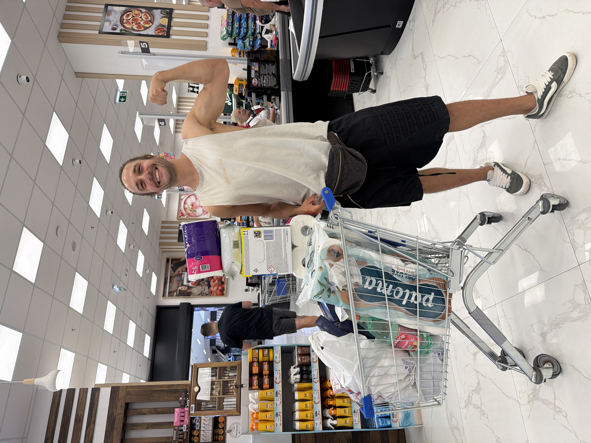 Here is a detailed description of the image:
**Scene:** The image is an interior shot, likely taken in a supermarket. The floor is polished white tile. The supermarket appears to be well-lit with ceiling-mounted lights.
**People:**
* **Subject:** A man is the main focus of the image. He is standing sideways to the camera, flexing his arm in a bicep pose. He is wearing a white tank top and dark shorts and Vans.
* **Other People:** Other patrons are seen in the aisles of the store, including one person in front of the subject.
**Objects:**
* **Shopping Cart:** The man is standing beside a shopping cart filled with groceries, including large packs of paper products and other items in white plastic bags.
* **Shelves:** Aisles of shelves are visible, stocked with various products, including bottled or packaged items, sunscreens and other items.
* **Store Fixtures:** Other store fixtures are present, like checkout counters and display units.
* **Signage:** There are signs hanging from the ceiling and mounted on the shelves.
**Actions:**
* The man is flexing his arm and smiling at the camera.
* People are walking in the aisles.
**Interactions:**
* The man poses for the camera.
**Location:** The text on the shelf below the cart reads "Budva". Therefore, the supermarket is located in Budva, Montenegro.