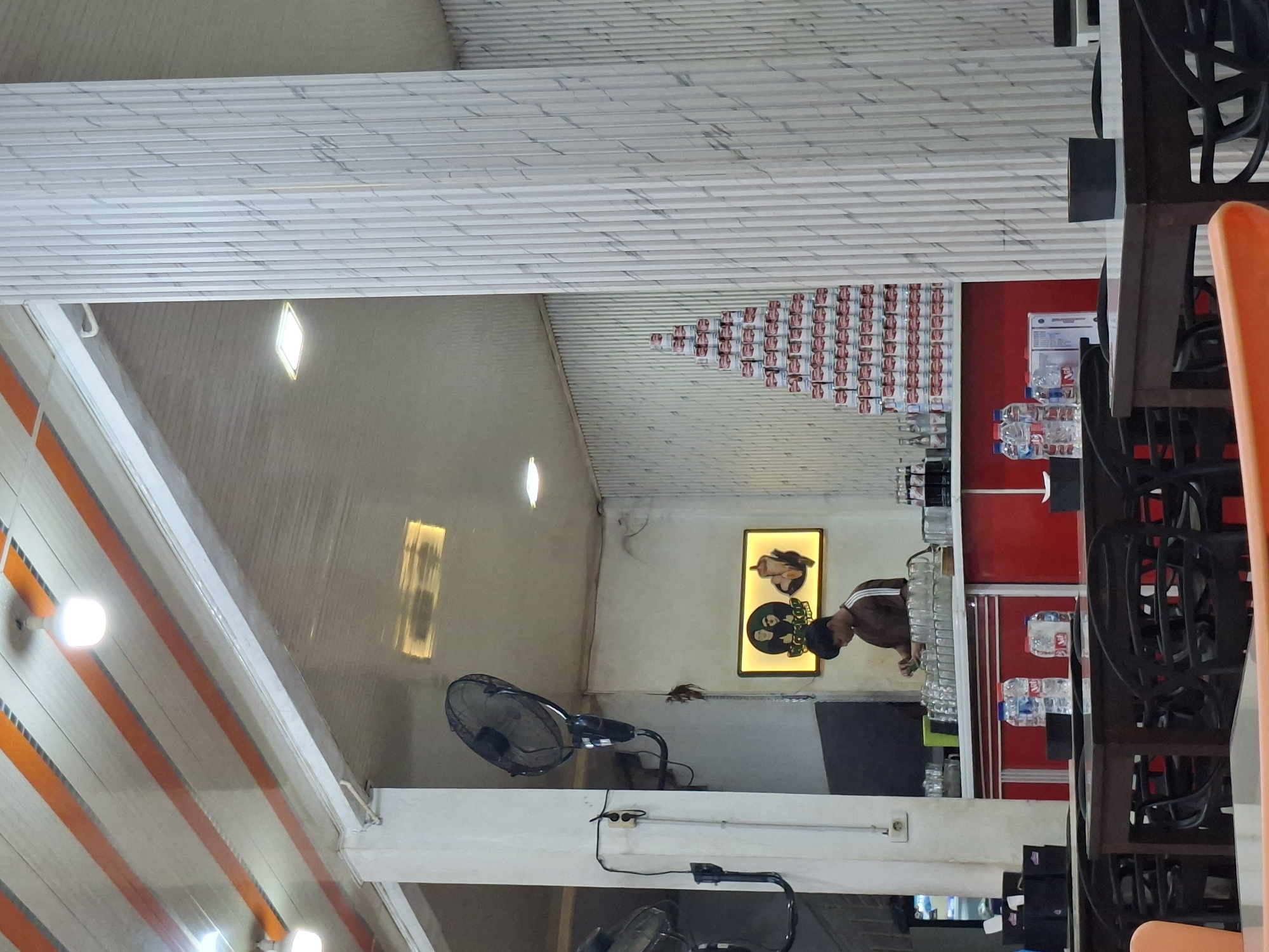 The image shows the interior of a cafe or restaurant in Sigli, Indonesia. In the foreground, there are several dark wooden tables with black chairs, suggesting a dining area. On one table, there are two plastic bottles of water. In the midground, a red counter is visible, with a young man in a brown shirt working behind it, surrounded by stacks of glasses and bottles. To the right of the counter, a tall, pyramid-like structure made of stacked cans is displayed. The walls of the establishment are decorated with a ribbed, textured pattern, and there are ceiling fans mounted on the white columns. A framed sign with cartoonish figures is also visible on the wall. The overall atmosphere appears to be casual and functional.