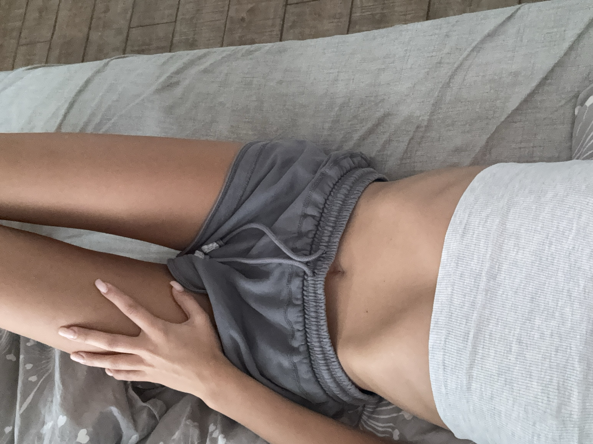 The image shows a close-up, overhead view of a person lying on a bed, with their legs extended upwards and their hand resting on their thigh. The person is wearing gray shorts and a light gray ribbed tank top. The bed is covered with a gray duvet featuring a subtle white heart pattern, and to the right, a section of wooden flooring is visible. The lighting suggests an indoor setting, likely a bedroom. The image does not provide any information or visual cues that could link it to the city of Chișinău, Moldova.