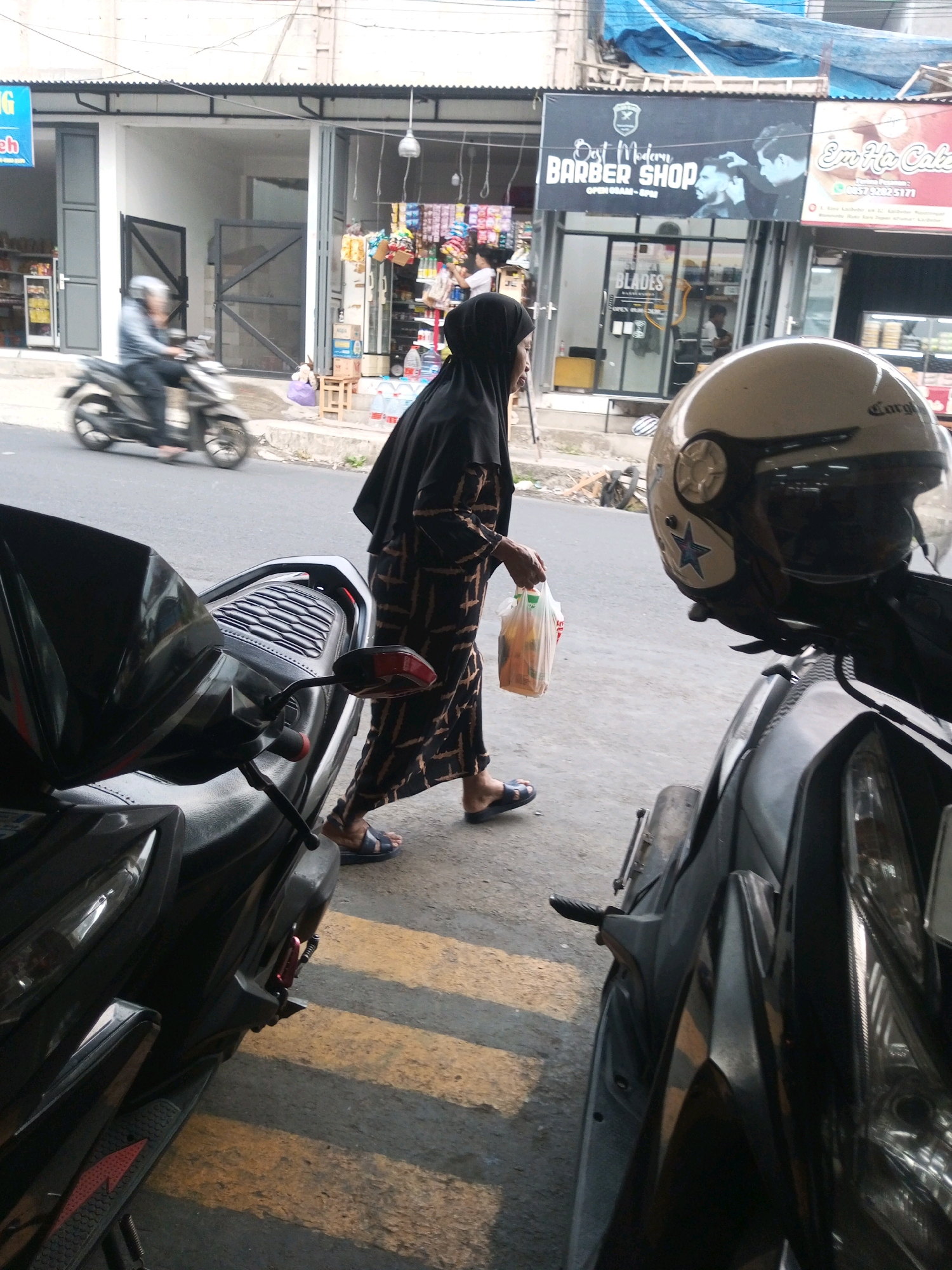 In Mojotengah, Indonesia, a woman wearing a black hijab and a patterned abaya walks on a sidewalk next to a street. She is carrying a transparent plastic bag filled with items. In the foreground, two motorcycles are parked. The motorcycle on the left is partially visible, while the motorcycle on the right has a white helmet with a blue star design placed on its handlebars. Behind the woman, a barber shop with signage that reads "Dost Modern Barber Shop" is visible. A person is riding a motorcycle in the background, and several storefronts line the street.