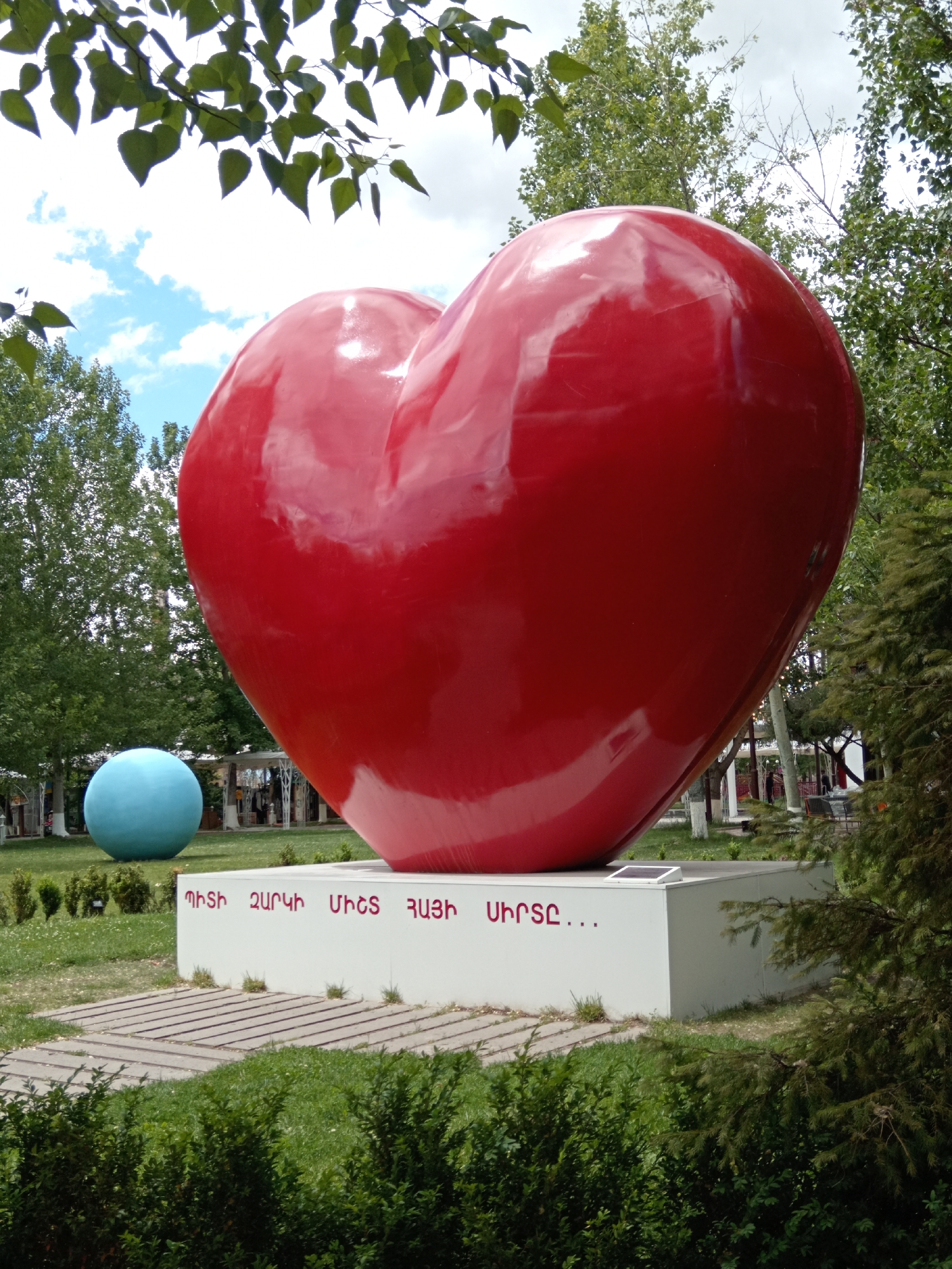 The image shows a large, shiny red heart sculpture on a white rectangular base, with text written in Armenian on the base. Behind the sculpture, there is a blue spherical object in a grassy field. The surrounding environment includes trees and greenery, suggesting a park or public space. The photograph appears to be taken outdoors during the daytime, with a partly cloudy sky visible. The location is in Yerevan, Armenia.