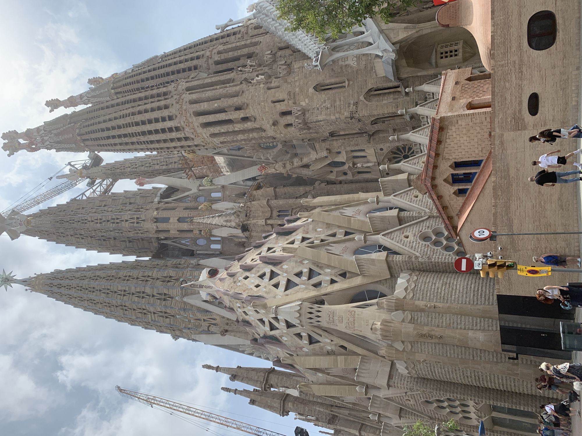 The image displays a bustling daytime scene at the iconic Sagrada Familia in Barcelona, Spain. The magnificent cathedral dominates the frame, showcasing its intricate, organic architecture with multiple towering spires, elaborate carvings, and detailed facades. The structure is constructed from light-colored stone, varying in shades of beige and grey, reflecting its ongoing construction with visible cranes integrated into the design.
The sky above is bright with scattered clouds, indicating clear weather. Sunlight illuminates sections of the cathedral, highlighting its complex textures and sculptural elements. Visible architectural details include various window shapes, including circular and rectangular openings, as well as geometric patterns and deep relief carvings. Latin and Catalan words are meticulously carved into the stone, including "SANDUS" (Sanctus), "EXCELSIS," "GLORIA," "OSANNA," "DOMINUS," "Pater," "Amen," "Credo," "Deus," "Meus," and "Plena de gràcies."
In the lower right portion of the image, several people are seen on a paved area adjacent to a simpler, reddish-brown brick building with small windows. These individuals, dressed in casual attire such as t-shirts, shorts, and jeans, appear to be tourists or passersby observing the grand structure. Their postures suggest a leisurely activity like sightseeing.
Nearby street furniture includes traffic lights, a red and white circular 'No Entry' sign, a speed limit sign displaying "10" (likely 10 km/h), and a yellow sign with a bicycle icon. A small tree with green leaves adds a touch of nature to the urban setting. The overall impression is one of a busy tourist destination, with the grand scale and detailed artistry of the Sagrada Familia being the focal point.