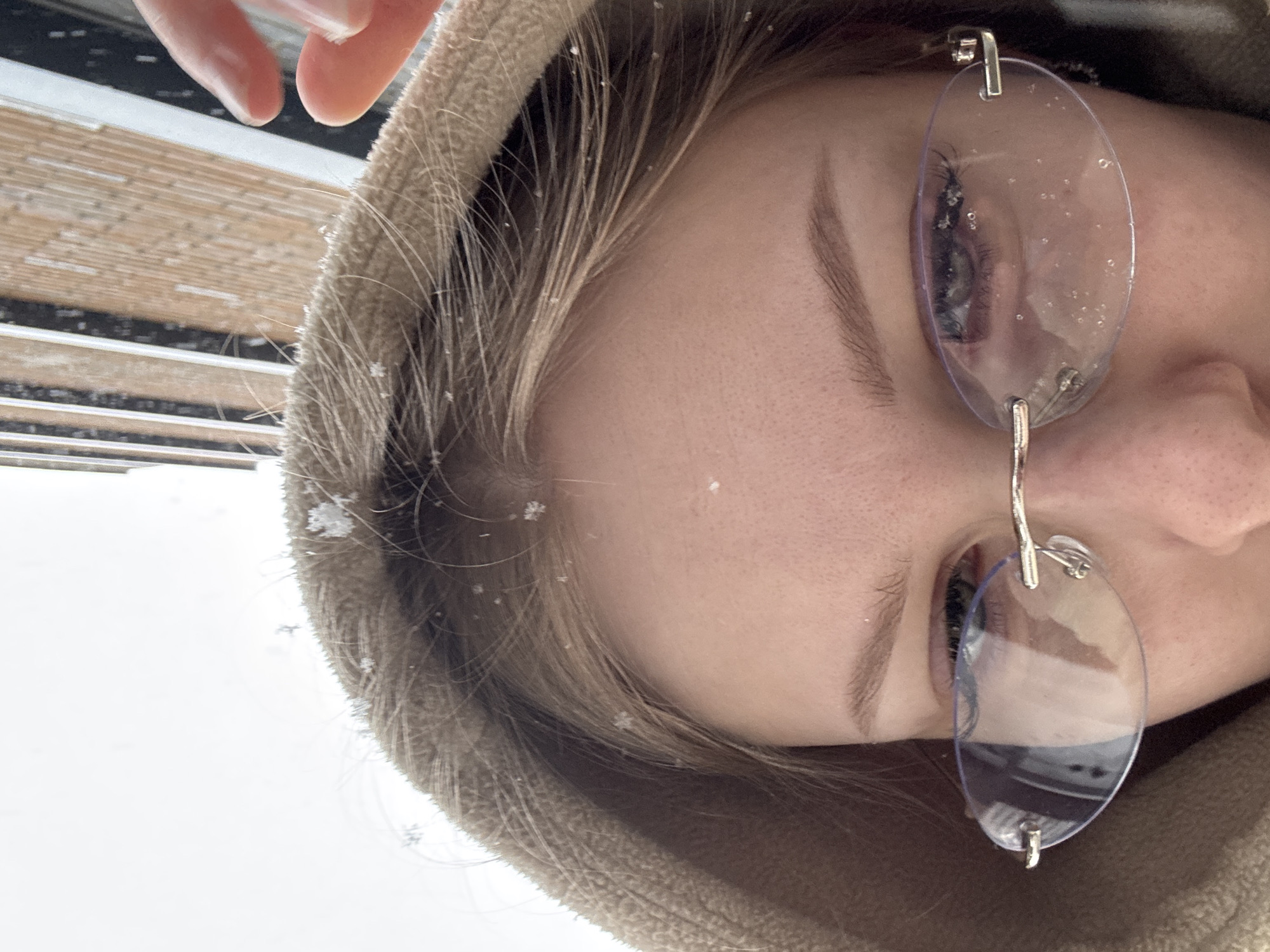 The close-up image features the upper right side of a person's face, tilted diagonally, focusing on their eye, eyebrow, forehead, and light-colored hair. The person is wearing a light brown or beige hooded garment, likely a jacket or sweatshirt, which frames their face.
Rimless glasses with clear, oval-shaped lenses are worn, and several small snowflakes are visible on the surface of the lenses. Numerous distinct snowflakes, some showing intricate crystalline structures, are also scattered across the person's hair and the textured fabric of the hood.
The scene takes place outdoors during snowy weather, evident from the visible snowflakes. The background is largely bright white, suggesting a snow-covered environment or an overcast winter sky. A blurry area with faint horizontal lines is visible in the top left, possibly indicating a building exterior or blinds. The lighting is bright and diffused, characteristic of an overcast daytime.
The activity appears to be simply experiencing the snowfall outdoors, perhaps looking up or taking a selfie. No specific emotions are discernible from the partial view of the face, and there is no visible text in the image. The provided location context is Perm, Russia.