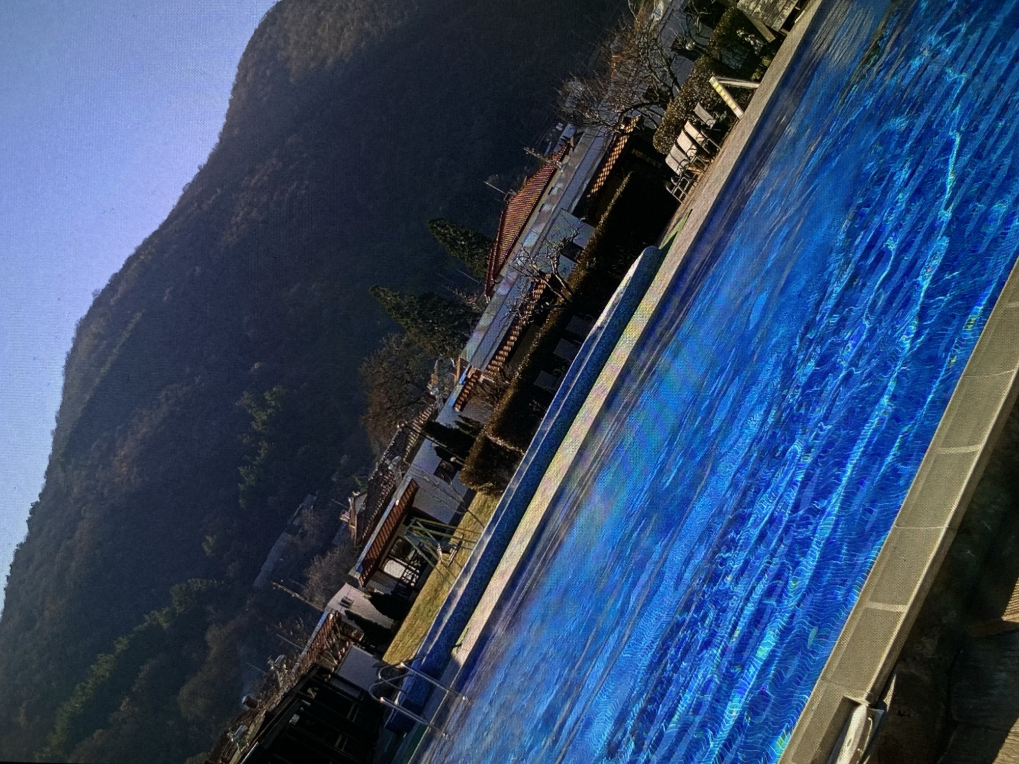 The image captures a vibrant blue outdoor swimming pool set against a mountainous backdrop in Bankya, Bulgaria. The shot is taken from a slightly elevated perspective, looking across the pool towards buildings and the mountains.
In the foreground, the large swimming pool dominates the right side of the frame, filled with clear, intensely blue water. Sunlight reflects off the surface, creating dynamic, shimmering patterns. The coping around the pool is a light greyish-brown. Towards the left end of the pool, chrome ladders provide access.
Beyond the pool, in the midground, several multi-story buildings are visible, likely part of a hotel or resort complex. They feature warm-toned, possibly red-tiled roofs and balconies. Between the pool and the buildings, there's a strip of green grass and some manicured bushes. Bare-branched trees, suggesting late autumn, winter, or early spring, are scattered around the property. Outdoor lounge chairs are arranged near the pool's edge, implying a leisure setting.
The background is dominated by a large, imposing mountain covered in dark, dense vegetation, possibly deciduous trees in their dormant state or evergreens. The mountain slopes upwards and diagonally across the upper left portion of the image, meeting a clear, bright blue sky.
The scene depicts a peaceful, sunny daytime setting, devoid of people, highlighting the natural beauty of the mountain landscape and the inviting ambiance of the resort. There is no visible text in the image.