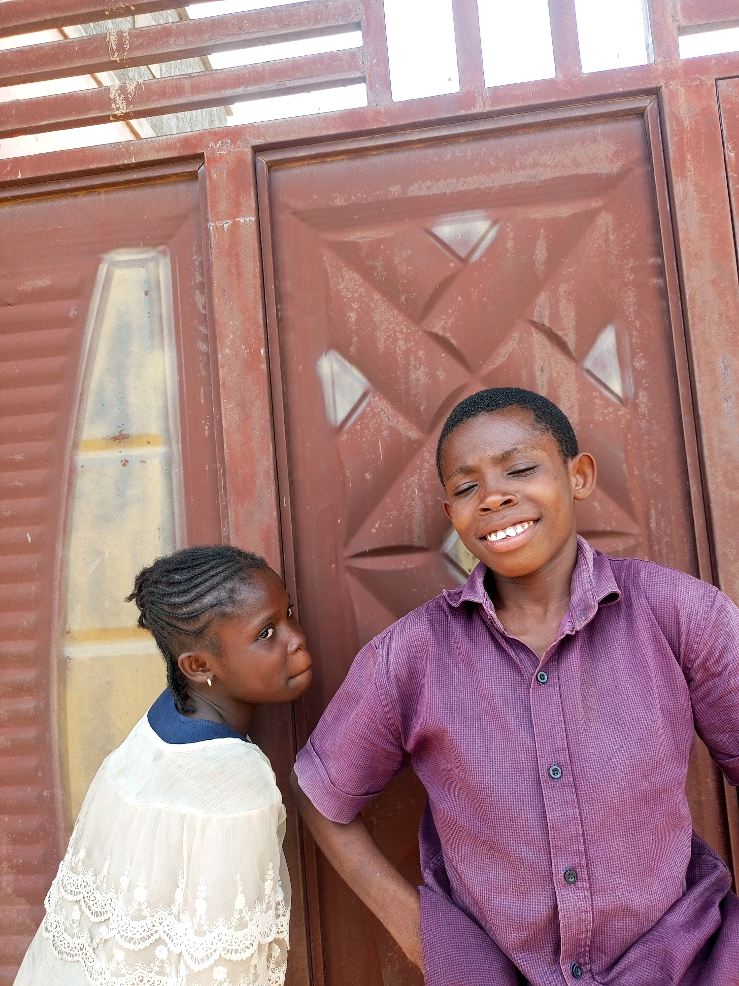 In Tempure, Nigeria, two young individuals stand before a weathered, reddish-brown metal gate. The gate features a diamond-patterned design and horizontal bars at the top. To the left, a young girl with braided hair is looking off to her right, wearing a white, lacy blouse with a dark blue collar. To her right, a young boy smiles broadly with his eyes closed, dressed in a purple, collared shirt. The lighting suggests an outdoor setting, possibly during the day.