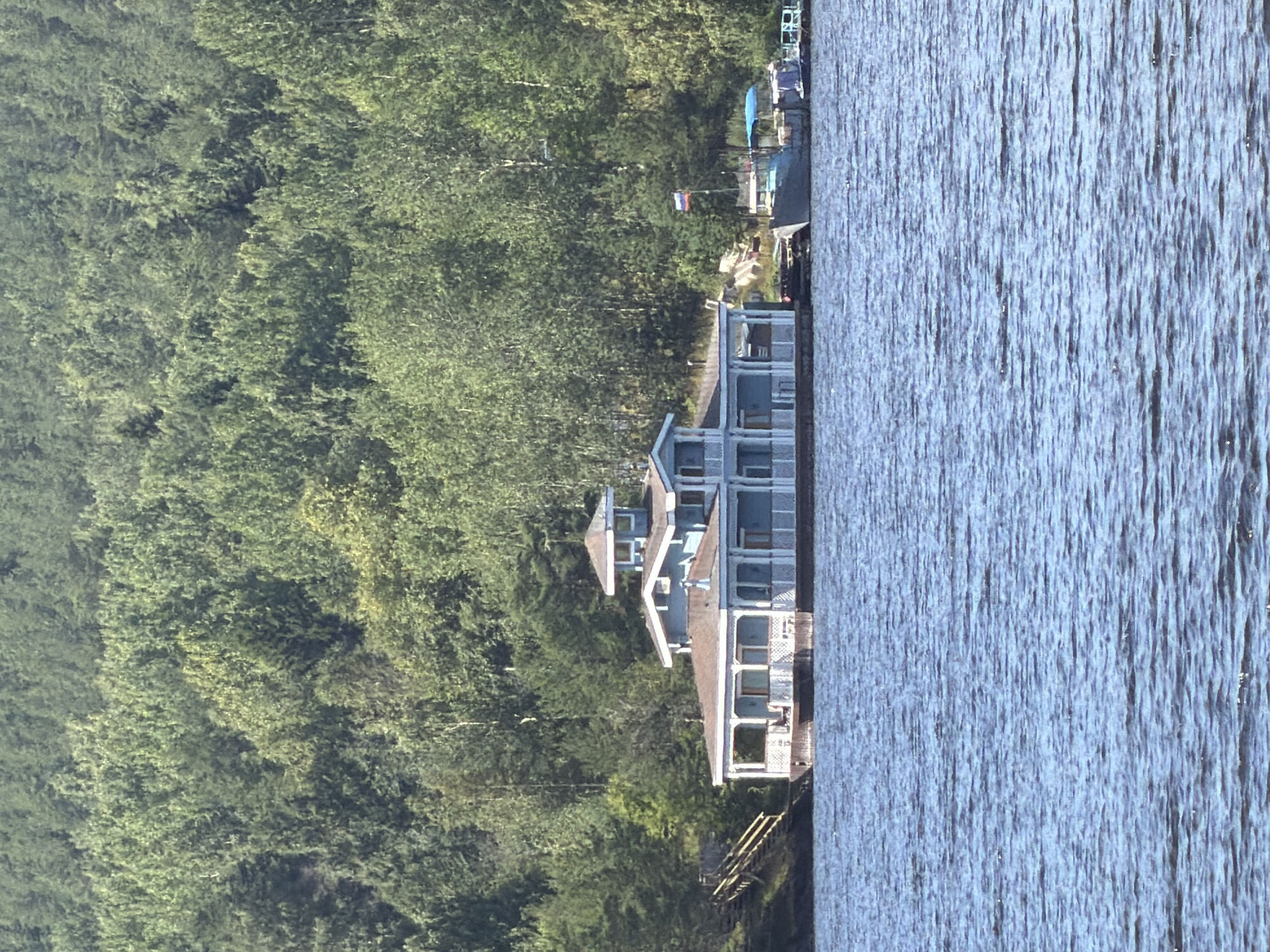 The image depicts a light blue, multi-story house situated on the water's edge, backed by a dense forest. The house features a wraparound porch with white pillars and railings, a dark-colored roof, and several windows. A set of wooden stairs with yellow railings leads from the shore to the house. Two boats are docked near the house, one of which displays a Russian flag. The water in front of the house is rippling. The scene appears to be a residential property in Yalguba, Russia, likely on a lake or river, given the presence of boats and the wooded backdrop.