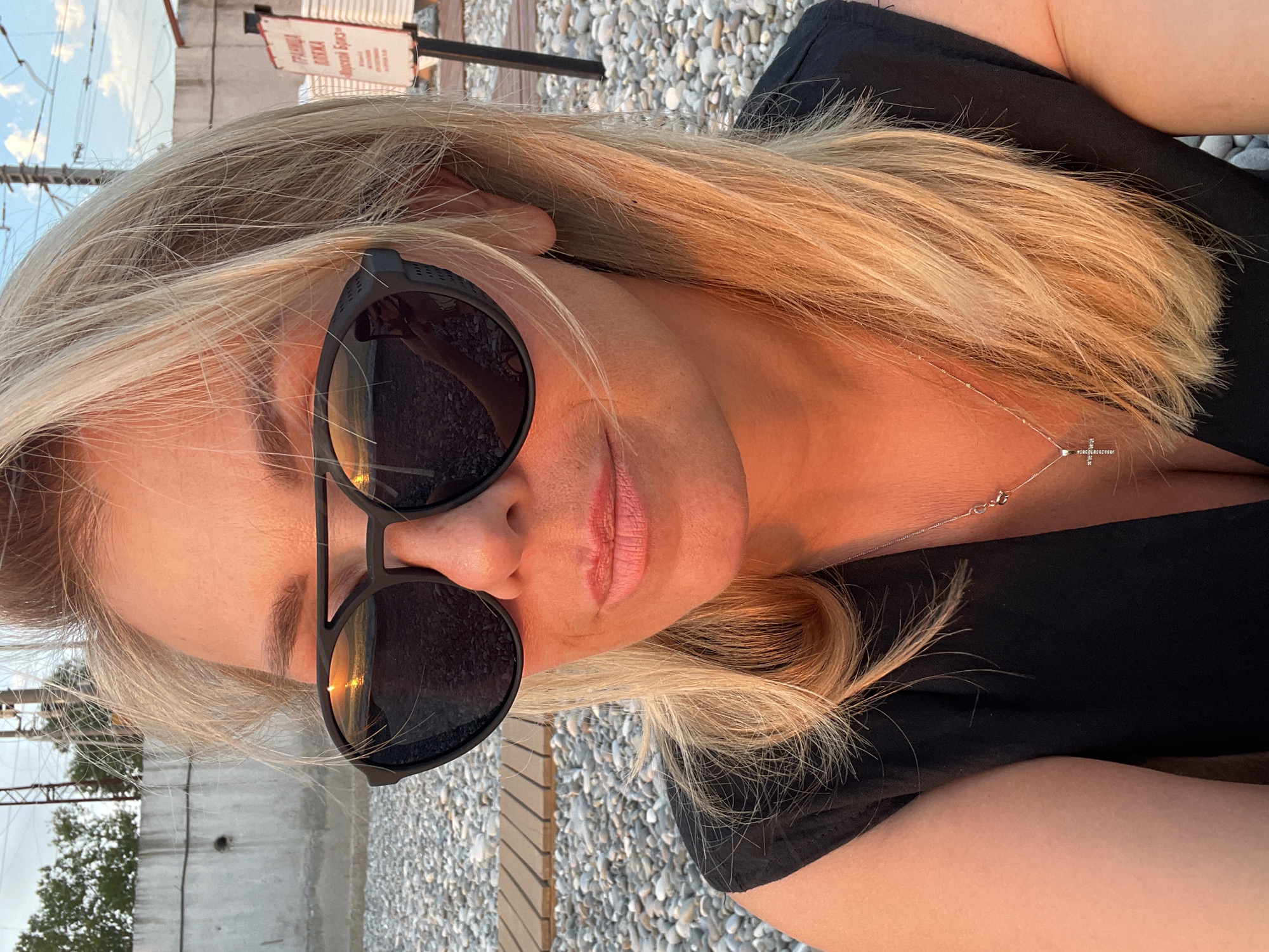 A woman with blonde hair and sunglasses is captured in a close-up selfie. She is wearing a black top and a silver necklace with a cross pendant. The background features a rocky ground, wooden seating, a concrete wall, and some industrial structures. The lighting suggests it's either sunrise or sunset, casting a warm glow on her face. There's a sign visible in the background with text in an unfamiliar language.