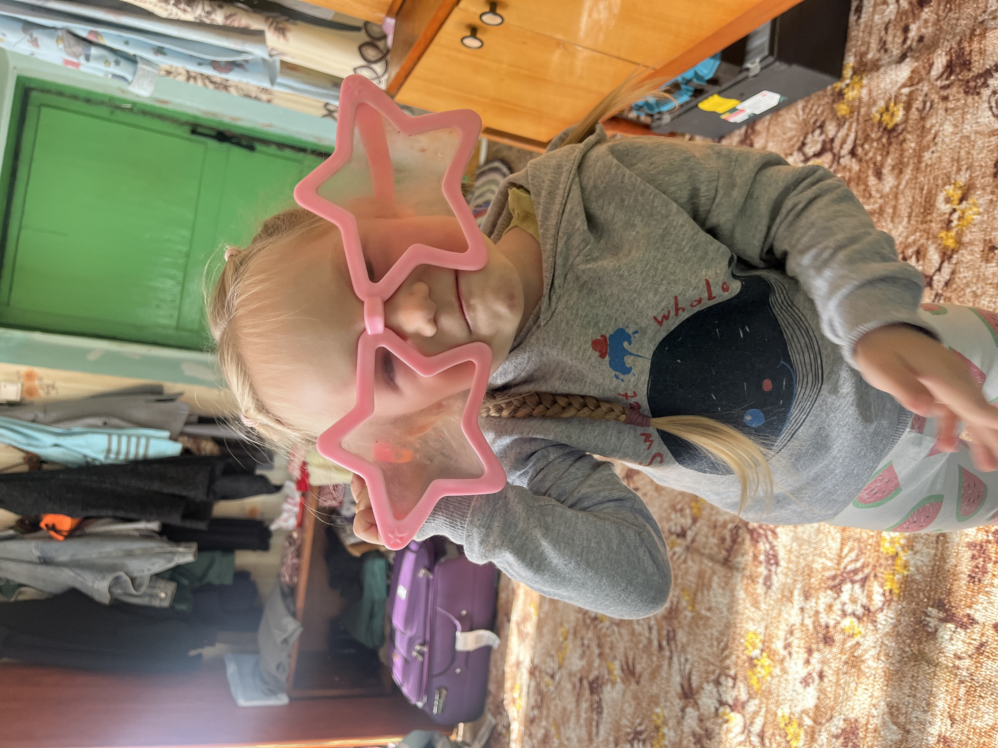 An outdoor scene in Kovriga, Russia shows a young girl in a gray hoodie and patterned pants wearing large pink star-shaped glasses. The girl is standing in a room with a patterned rug on the floor. To the left, there is a purple suitcase and a shelf with clothes. To the right, there is a wooden cabinet with drawers and a green door in the background. The lighting in the room is dim, with sunlight streaming in from the left.