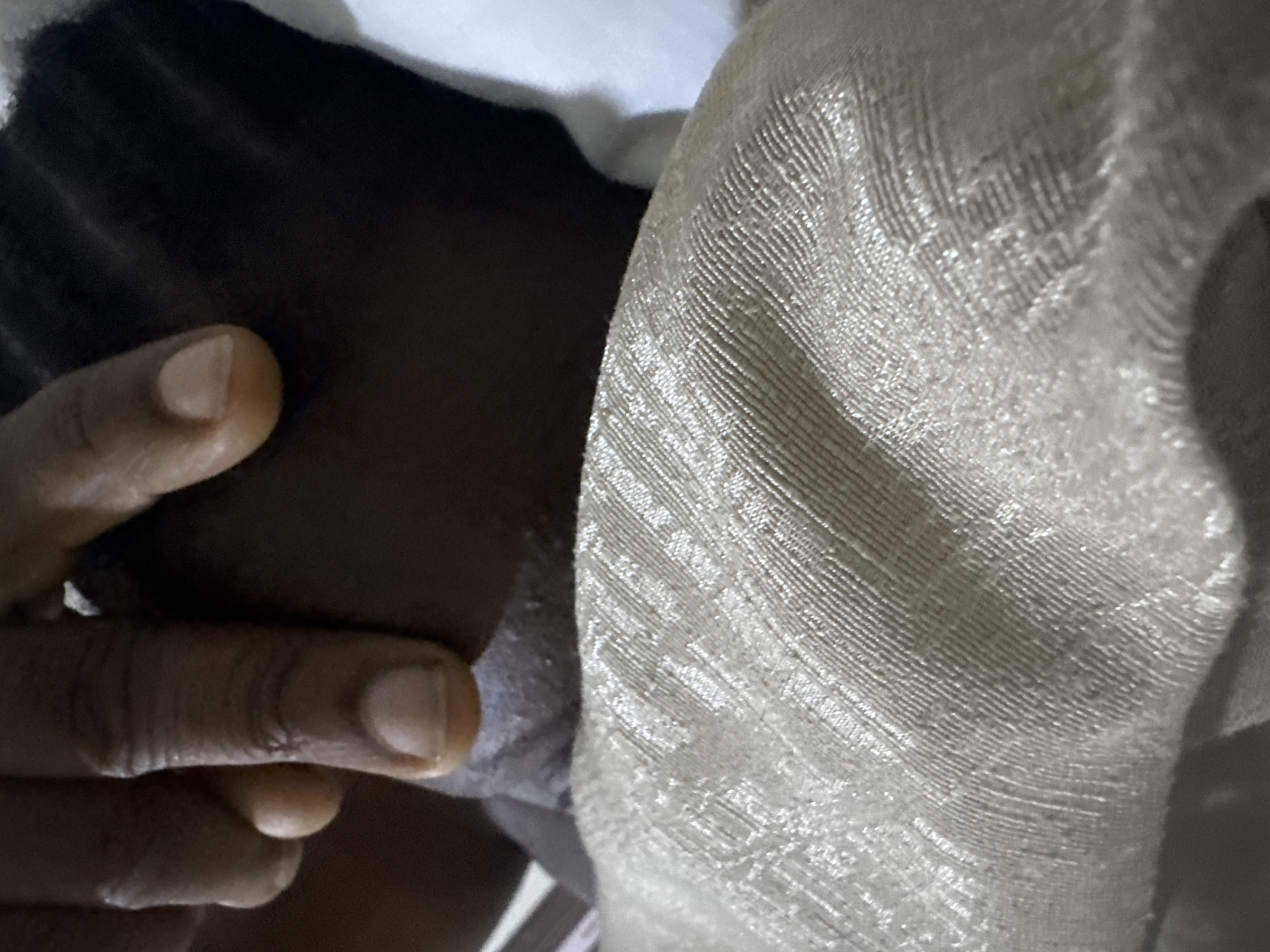 In Karu, Nigeria, a close-up shot captures a human hand gently touching the head of a person. The hand, with its dark skin tone and visible fingerprints, is positioned near the forehead and hair, suggesting a moment of care or attention. The person's head is partially obscured, but their dark hair is visible. A textured, light-colored fabric, possibly a pillow or blanket, occupies the foreground, creating a soft backdrop for the interaction. The overall lighting is subdued, with a slight sheen on the fabric.