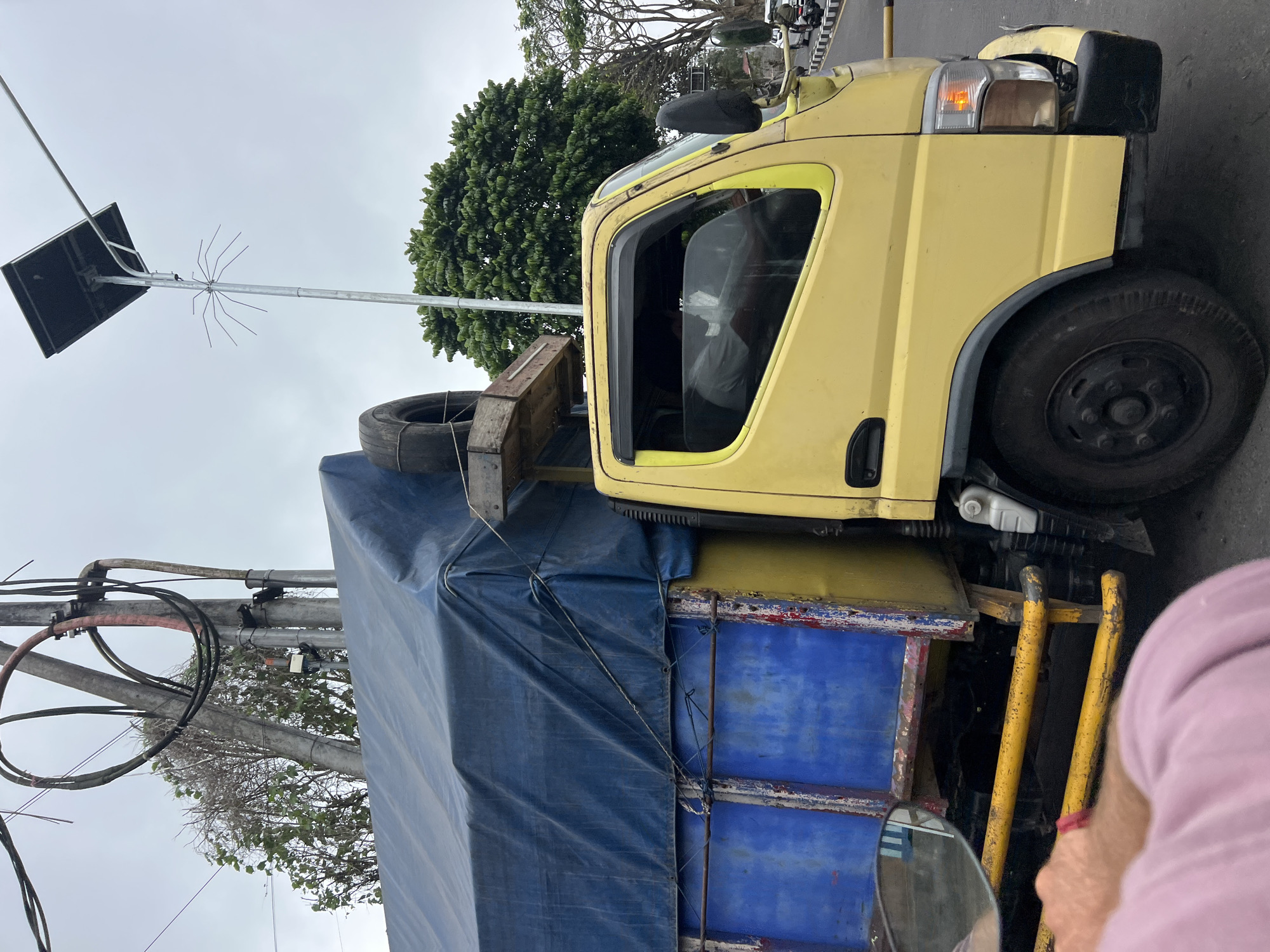 A yellow truck is seen from the side, driving on a paved road. The truck has a blue tarp covering its cargo bed, and a spare tire is secured on top. In the background, there are utility poles with wires, lush green trees, and a cloudy sky. A person is visible inside the truck's cab, appearing to be the driver. The scene is likely in Rendang, Indonesia, given the overall environment and the presence of typical Indonesian street elements.