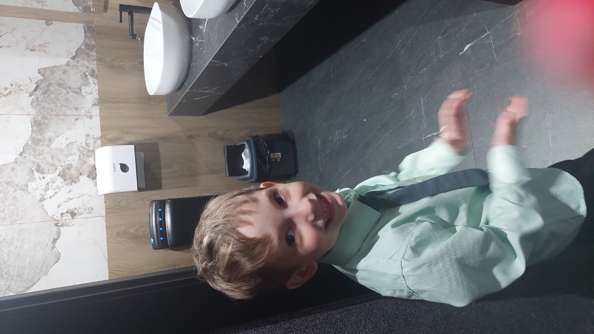 A young Caucasian boy with light brown hair and a blue tie is smiling at the camera. He is wearing a light green collared shirt and dark pants. He is standing in a modern public restroom. The walls are tiled with a marble-like pattern and wood panels. There is a white paper towel dispenser on the wall, and a dark gray trash can with a black liner is next to it. To the right, there is a dark countertop with two white sinks and a black faucet. The floor is dark gray tile. The boy's hands are blurred, indicating movement. The image appears to have been taken in Komsomolsk-on-Amur, Russia.