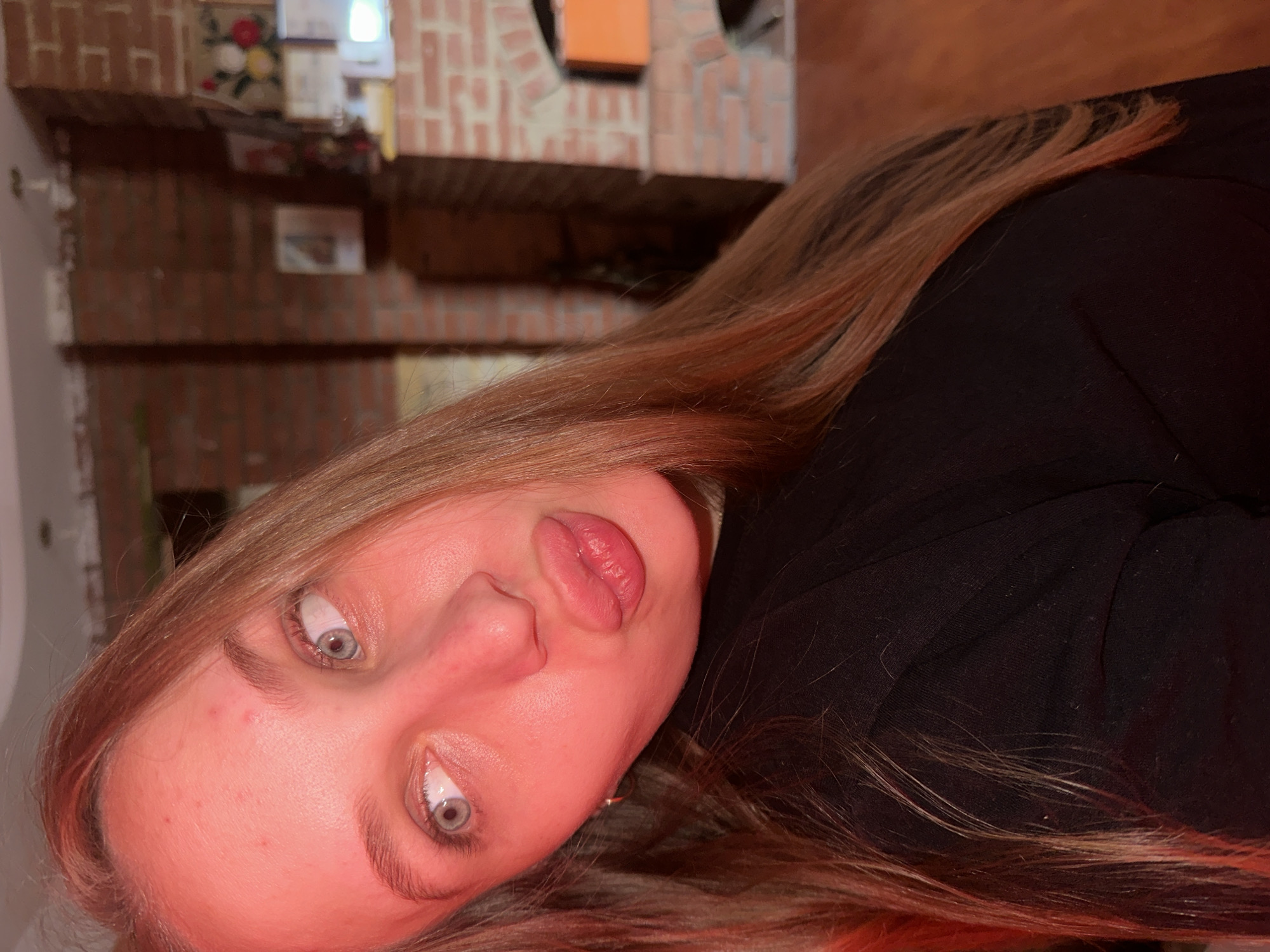 This image depicts a young adult female in an indoor setting, located in the city of Chaikovsky, Russia. The subject is positioned facing forward with her head tilted to the viewer's left. She has long, straight, light brown hair, parted on the side, falling over her right shoulder. Her eyes are light blue/grey and gaze towards the upper right of the frame (viewer's left). Her lips are tightly pursed into an exaggerated 'duck face' or 'fish face' expression, with a slight gleam on the lower lip. Small blemishes are visible on her forehead. She wears a dark, likely black, long-sleeved top. A small metallic earring is visible on her left earlobe. The scene is heavily illuminated by a strong reddish-orange light source, casting a saturated, warm hue over the subject and surroundings. The background features exposed reddish-brown brick walls, including a large brick structure resembling a fireplace or oven with an arched opening on the right side of the frame. Various decorative items, including framed pictures and what appears to be embroidered artwork with floral patterns, are visible on the brickwork and a mantelpiece. A light-colored wooden floor is partially visible in the lower right background. A portion of a white ceiling with a small fixture is visible at the top.