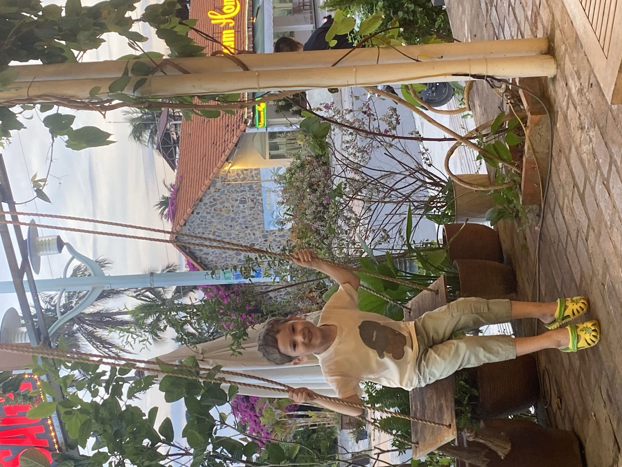 A child is sitting on a swing suspended by rope, surrounded by lush vegetation, likely in the city of Phu Thuy Ward, Vietnam. The child wears a light-colored t-shirt with a brown bear design, light green pants, and bright yellow and green sandals. The background reveals building structures, including a section with a red tile roof and a stone wall, interspersed with greenery and vines. The vegetation is abundant, with flowers and leafy plants visible throughout.