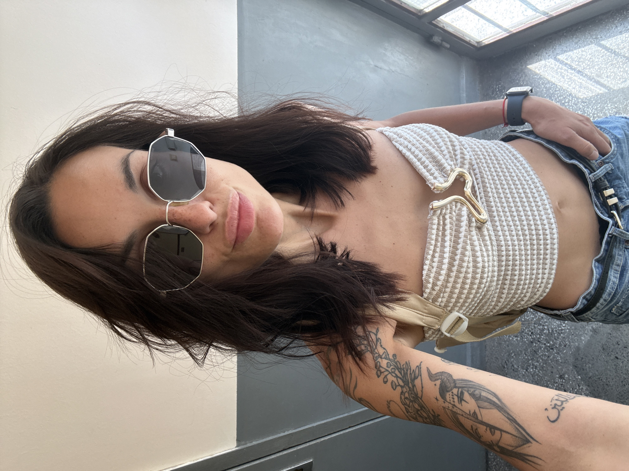 A woman with dark hair, wearing sunglasses, a cropped knit top, and jeans, poses for a selfie. She has tattoos on her left arm. She is standing indoors, with a door and a window visible in the background. The floor appears to be made of concrete or stone tiles. The wall behind her is painted a light color, with a gray section above it. The overall environment suggests an urban setting, possibly an apartment building or an office. There is no specific information available to confirm this image was taken in Pleven, Bulgaria.