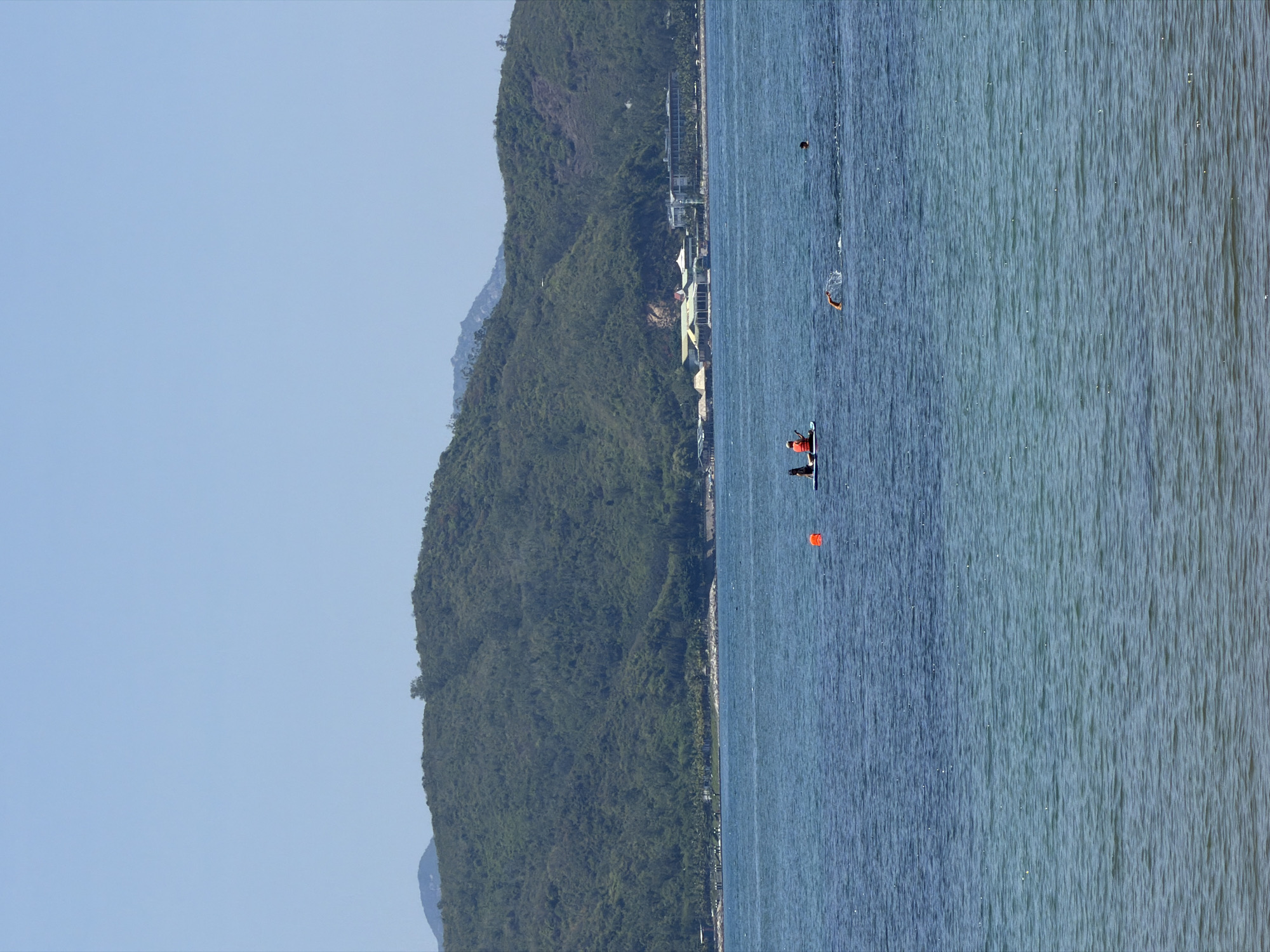 This image captures a coastal scene in Bac Nha Trang Ward, Vietnam, under a clear, light-blue sky. A large, tree-covered mountain range forms the background, extending across the horizon. At the base of these mountains, along the shoreline, several buildings are visible, including structures with light-colored roofs, resort-style architecture, and a pier on the left. The foreground features calm ocean water, displaying a gradient from deep blue to lighter blue-green. In the water, two individuals are seated on a light-colored stand-up paddleboard; one wears an orange life vest, and the other wears dark clothing. An orange spherical buoy floats to the right of the paddleboard. To the right of the buoy, a person is actively swimming freestyle, with one arm out of the water creating a splash. Further to the right, another individual's head is visible above the water surface.