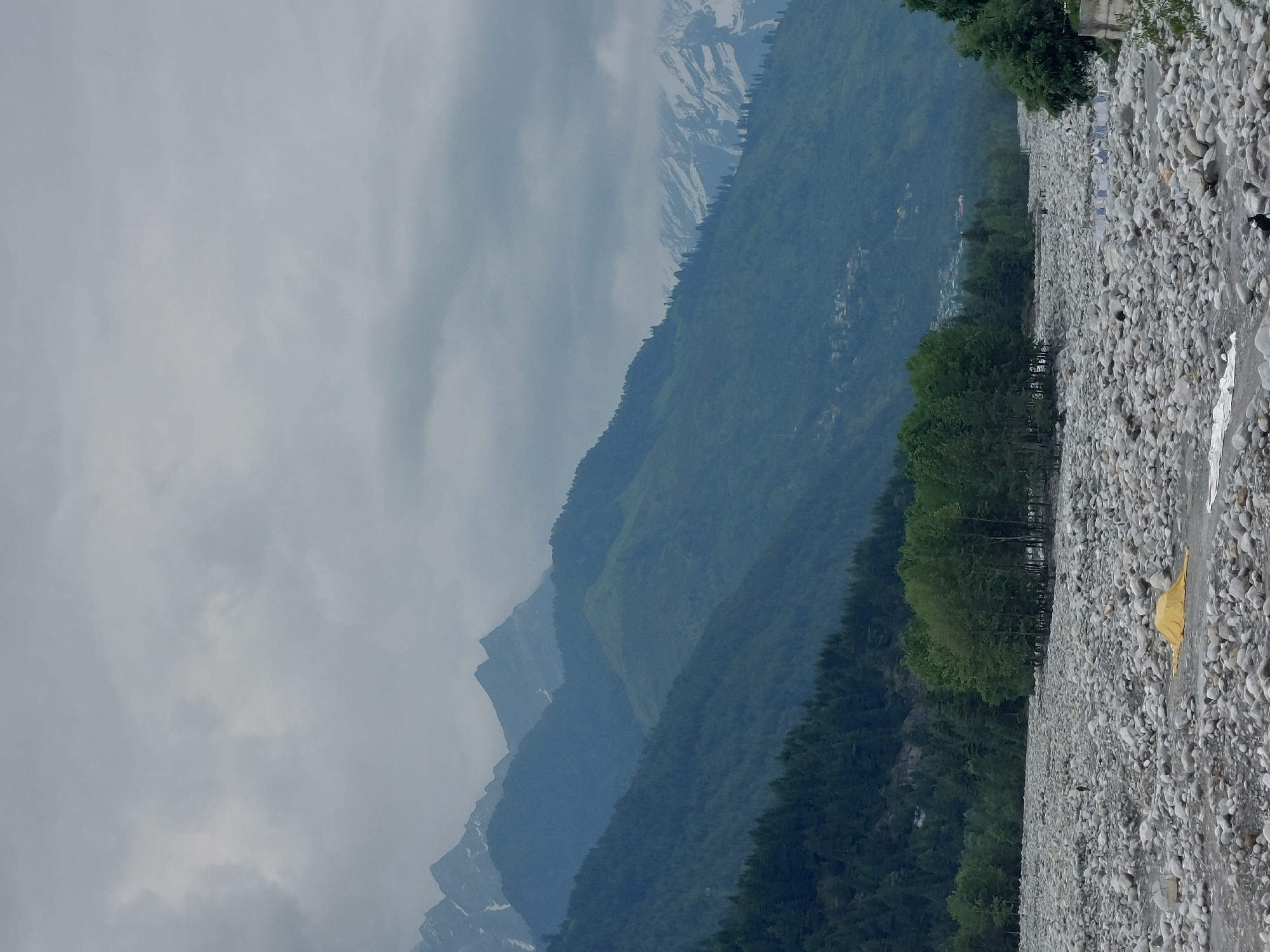The image depicts a wide, rocky riverbed in Vashisht, India, with a backdrop of majestic mountains. The riverbed is densely covered with smooth, gray stones of various sizes. In the foreground, a yellow tarp is spread out on the rocks. Further into the riverbed, several individuals are visible, some appearing to be walking or standing among the stones, and others potentially engaged in activities like washing or drying clothes, indicated by lines of fabric. A cluster of green trees forms a prominent line along the edge of the riverbed, separating it from the steep, forested slopes of the mountains. The mountains themselves are layered, with darker green forests covering the lower inclines and lighter, possibly snow-dusted peaks visible in the distance under a cloudy, overcast sky. The overall scene conveys a sense of natural grandeur and a tranquil, rugged landscape.