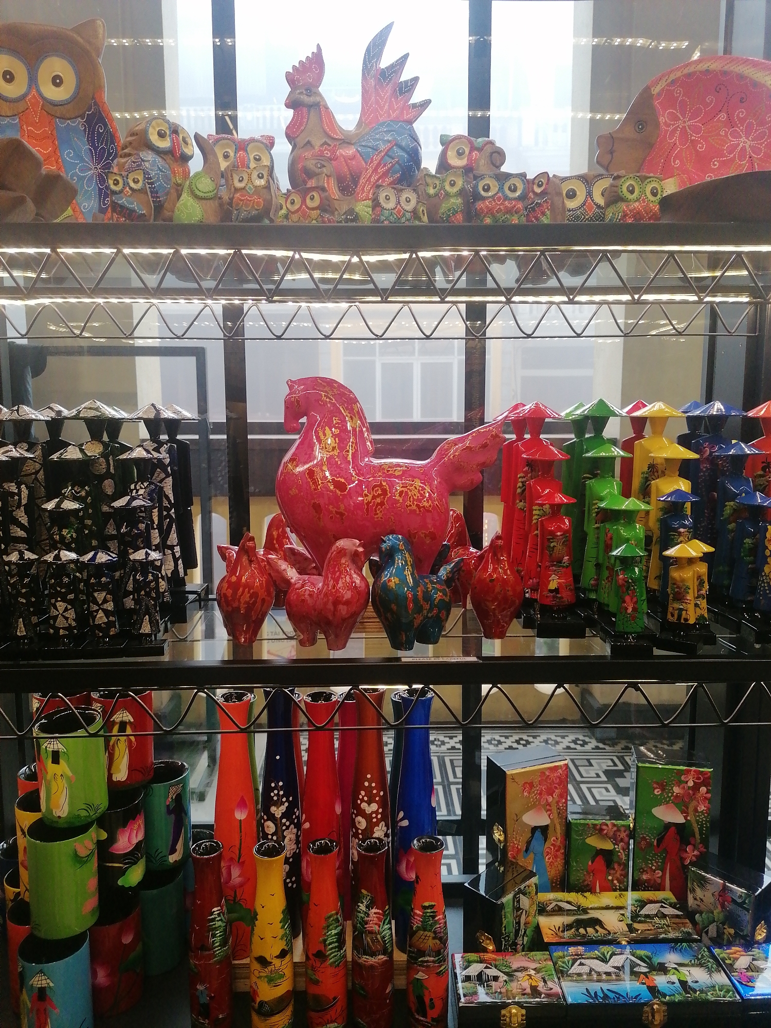 This image captures a vibrant display of handicrafts and decorative items arranged on multi-tiered shelves within what appears to be a souvenir shop or market stall. The setting is indoors, with artificial lighting illuminating the products.
The top shelf showcases a collection of brightly painted wooden animal figurines. Prominently featured are several owls of varying sizes, adorned with intricate patterns in blue, orange, yellow, and green. A large wooden rooster, similarly decorated with red and blue hues, stands among them. To the right, a large, stylized wooden fish or pig sculpture, painted red with white floral patterns, is also visible.
The middle shelf features a series of tall, slender figurines resembling people wearing traditional Vietnamese conical hats (nón lá). These are arranged in rows and come in various colors, including black-and-white patterns, red, green, yellow, and blue, some with painted scenes on their bodies. Beside them is a large, glossy hot pink sculpture of a horse, embellished with gold flecks, and smaller matching red and blue bird-like figures. A small white label with the text "PLEASE BE CAREFUL" is faintly visible on this shelf.
The bottom shelf holds a diverse array of items. On the left are numerous cylindrical objects, possibly mugs or pen holders, in green, red, black, and blue, decorated with scenes of people in conical hats and lotus flowers. To their right are several tall, narrow vases, predominantly red, blue, yellow, and orange, adorned with landscapes featuring traditional Vietnamese houses, boats, and figures, as well as floral motifs. Further right, an assortment of rectangular boxes or plaques, some standing upright and others laid flat, display colorful painted scenes. These depict people in conical hats, pastoral landscapes with houses, trees, animals (like a tiger or buffalo), and boats on water. The boxes have visible brass-colored clasps.
The overall scene suggests a lively collection of local artisanal products, indicative of Vietnamese culture, arranged for viewing and sale.