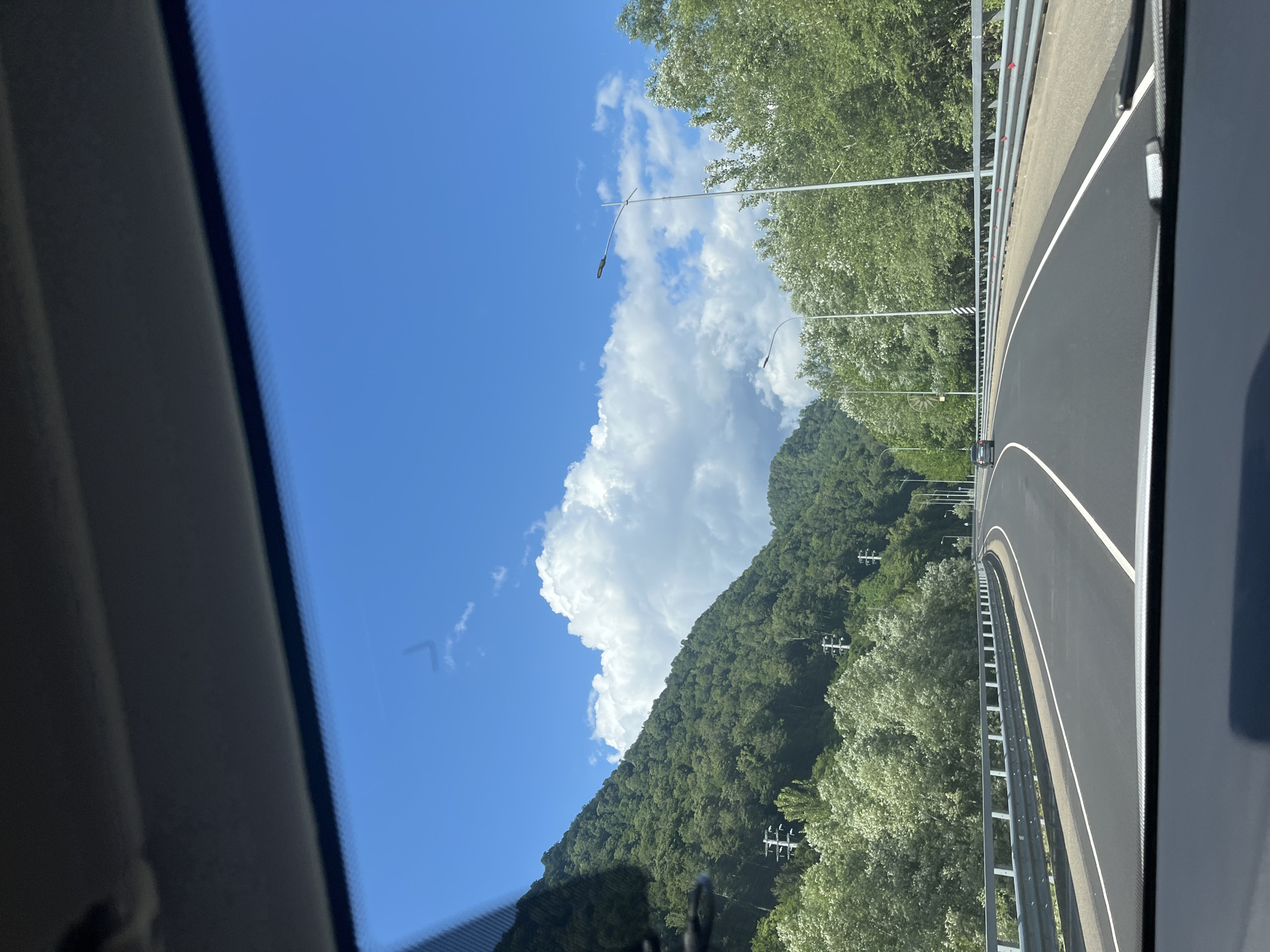 This image depicts a view from the interior of a vehicle looking out at a road curving through a mountainous, forested area. The sky is bright blue with scattered white cumulus clouds. On the left side of the road, a steep, densely forested mountain rises. On the right side, there is a line of trees and then more forested mountains in the distance. The road itself is asphalt, with clear white lane markings and a metal guardrail along the side. There are streetlights visible along the road, and power lines and poles can be seen on the mountainside. A few cars are visible on the road in the distance. The lower portion of the image shows the dashboard and windshield frame of the vehicle from which the photo was taken. The scene is likely a highway or major road in a rural, mountainous region. The location is identified as Kepsha, Russia.
