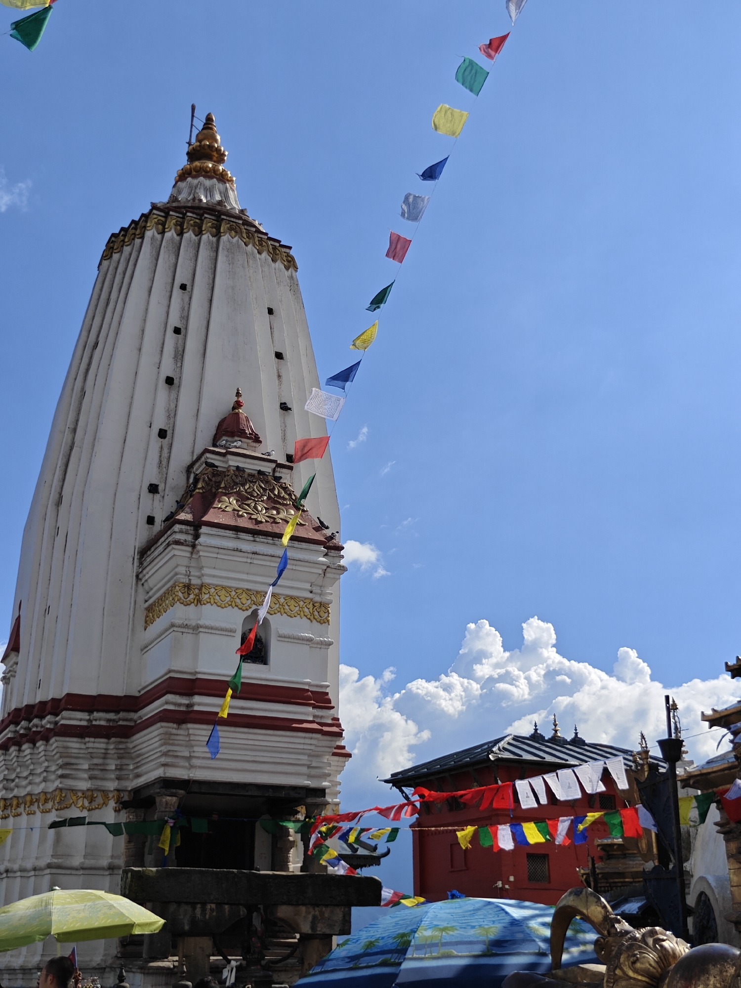 The image depicts the Swayambhunath Stupa complex in Kathmandu Metropolitan City, Nepal, on a clear, sunny day with scattered clouds. The main subject is the white, multi-tiered stupa, adorned with golden finials. Prayer flags in various colors (blue, white, red, green, yellow) are strung across the scene, fluttering in the breeze, some trailing from the stupa and others forming lines against the sky.
In the mid-ground, a red-roofed building with decorative eaves is visible, also decorated with strings of prayer flags. Various architectural elements of the complex, including golden decorative accents and intricate stonework, are discernible. In the foreground, a partially visible yellow umbrella and part of a blue umbrella with palm tree motifs are present, suggesting a public area or marketplace. A person's shoulder is visible near the yellow umbrella. The overall scene conveys a sense of cultural and religious significance, set against a backdrop of a bright blue sky.