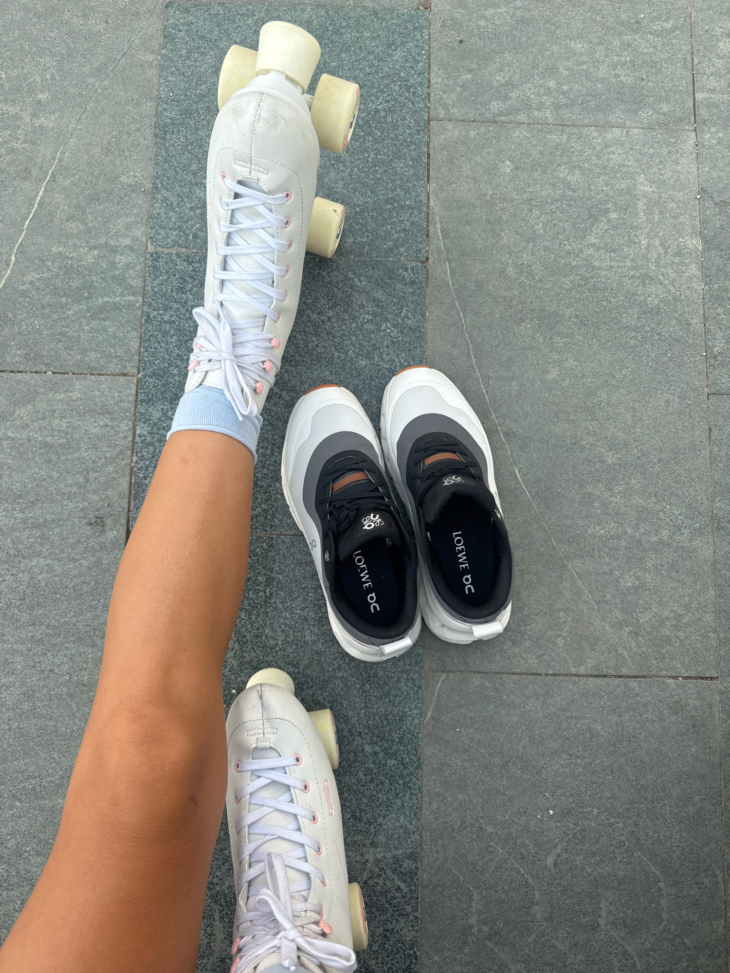 An overhead shot shows a person's left leg and right foot on a grey tiled surface. The leg is wearing a light blue sock and a white roller skate. The roller skate is laced with white laces and has pink eyelets. The person's right foot is wearing a white and grey sneaker with black laces. The sneaker has the text "LOEWE OC" on the insole. The tiled surface appears to be outdoors, possibly in a public space.