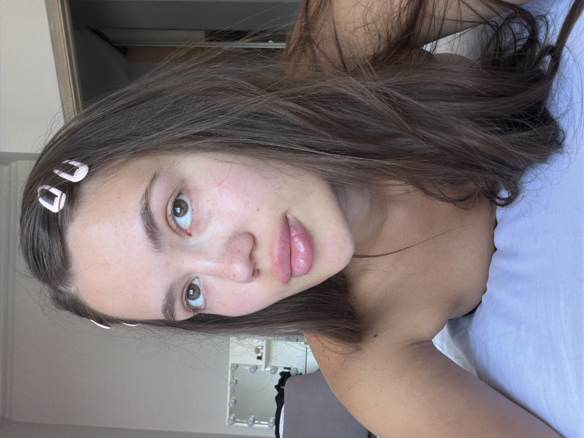 A young woman with brown hair and light skin is lying on a white bed. She is looking up and to the right. Her hair is parted in the middle and she has two pink barrettes in her hair on the left side. She has freckles on her cheeks and nose. Her lips are full and shiny, and appear to be wearing lip gloss. She is wearing a light-colored top that is off her shoulders, exposing her arms and upper chest. Behind her, a vanity mirror with lights is visible, as well as a closet door. The image is taken from a close-up perspective, focusing on the woman's face and upper body. The lighting is natural and bright, suggesting it is daytime.