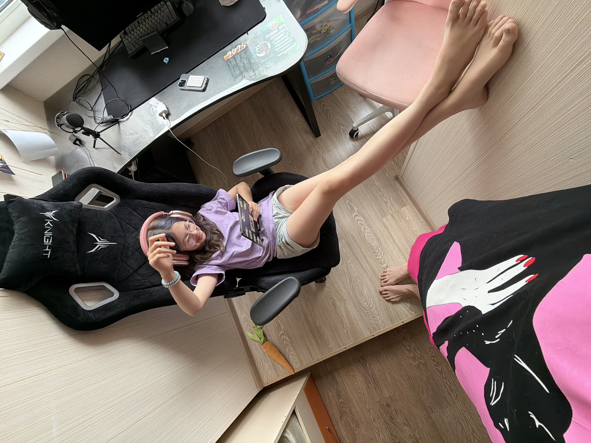 A young girl with headphones and glasses is sitting in a black gaming chair, holding a tablet. She is wearing a purple t-shirt and grey shorts. A stuffed carrot is on the floor next to the chair. The scene appears to be an indoor setting, possibly a bedroom or a gaming room, with a desk, computer monitor, and other gaming accessories visible. The floor is wooden and there is a pink chair in the foreground. The image does not contain any visual cues that would indicate its location in Birobidzhan, Russia.