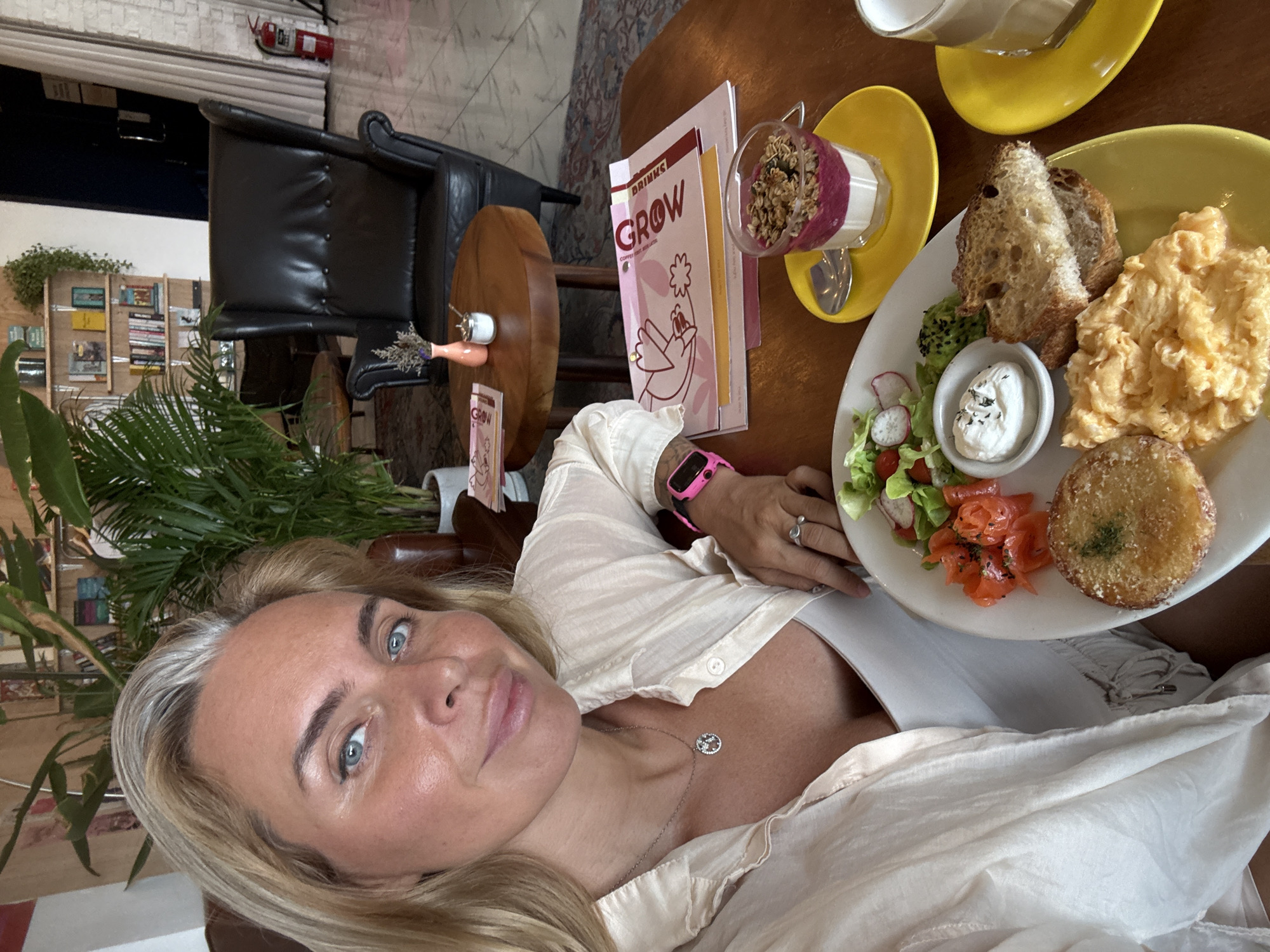 In a brightly lit indoor setting, possibly a cafe in Baan Bo Phut, Thailand, a woman with blonde hair and blue eyes is taking a selfie. She is wearing a light-colored, open collared shirt over a white tank top, and a silver pendant necklace. A bright pink smartwatch is visible on her left wrist. In front of her, on a wooden table, is a plate of food containing scrambled eggs, toasted bread, smoked salmon, a small salad with radishes and lettuce, and a circular fried item that appears to be a potato cake. Beside the plate, on a yellow saucer, is a glass of yogurt parfait with granola. Another yellow saucer with a glass containing a beverage is to the right. In the background, a large leather armchair is positioned near a wooden table holding a small vase with dried flowers and a sugar dispenser. A bookshelf filled with books is visible further back, along with lush green plants and a fire extinguisher.