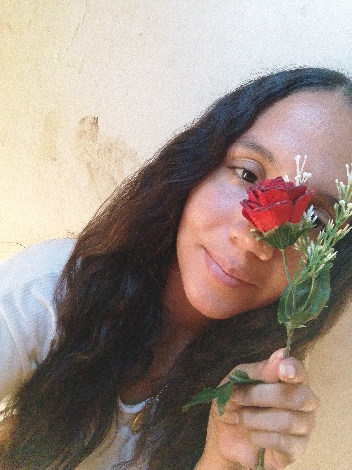 A woman is holding a red rose with green leaves and small white flowers in front of her face. She is smiling slightly, looking directly at the camera. Her hair is long, dark, and wavy. She is wearing a white shirt. The background is a textured, off-white wall. There is no indication of the city or location of Juan Griego, Venezuela.