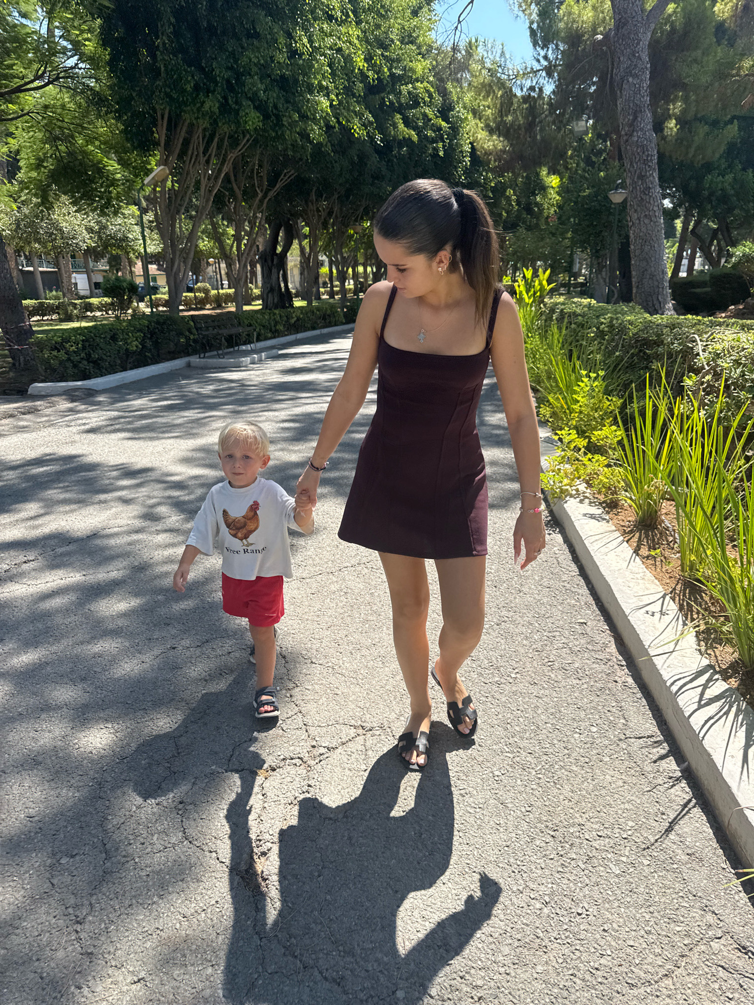 A woman and a young child are walking on a paved pathway in a park. The woman is wearing a dark plum colored dress and black sandals. She is holding the child's hand. The child is wearing a white t-shirt with a chicken graphic and red shorts, and sandals. The pathway is shaded by trees, and there are green bushes and trees in the background. The image was taken on a sunny day. This scene is consistent with locations like the Limassol Municipal Gardens in Limassol, Cyprus.