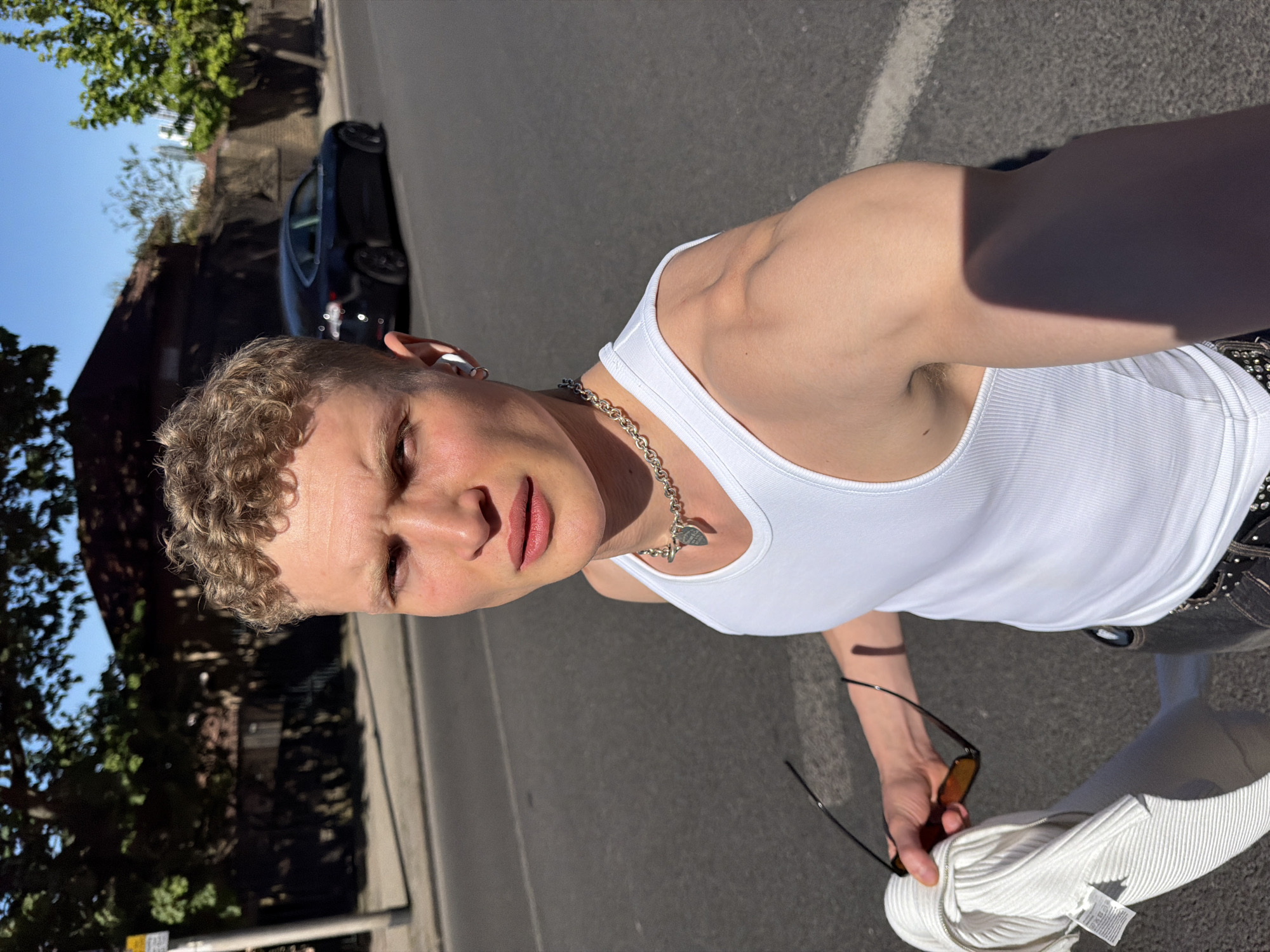 A young man with light, curly hair is depicted in a brightly lit outdoor selfie. He wears a white ribbed tank top and a silver chain necklace featuring a heart-shaped pendant, possibly from the "Return to Tiffany" collection. White wireless earbuds are visible in his ears. He holds dark sunglasses with amber lenses in his right hand, along with a white, ribbed zippered garment that has a tag attached. A dark belt with metal embellishments is visible around his waist.
The man looks directly at the camera with a neutral expression. Strong sunlight illuminates the scene, casting prominent shadows, such as from his arm onto his side. The background features an urban street or sidewalk under a clear blue sky, with a dark car parked on the side of the road. Green trees and parts of buildings, some with brick facades, are visible further in the distance. The ground is paved asphalt. The scene suggests a casual daytime setting in an urban environment, consistent with London, United Kingdom. No specific text is clearly legible on the clothing tag or in the background.