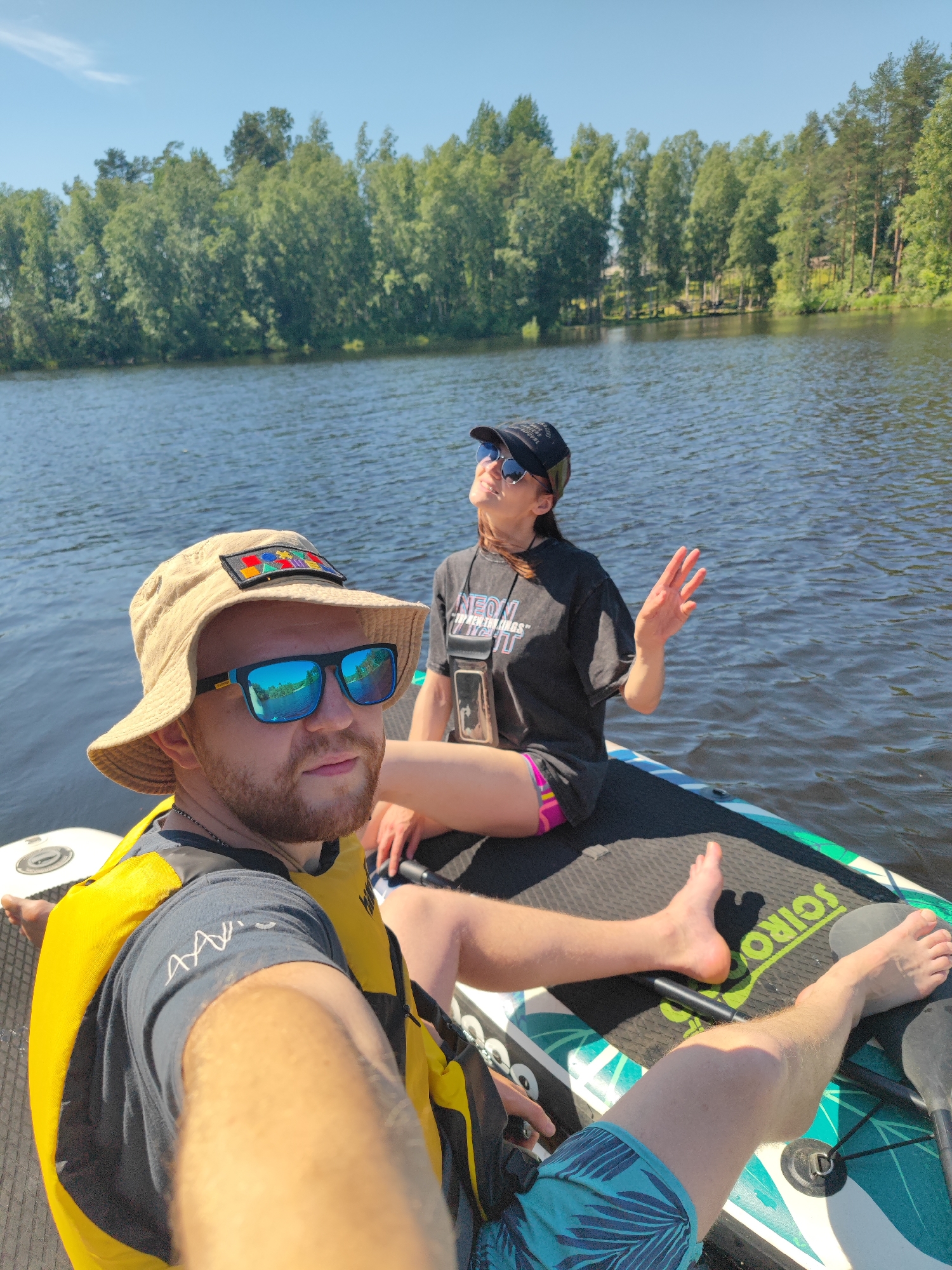 Two individuals are on a paddleboard on a calm body of water, surrounded by lush green trees. The person in the foreground, wearing a bucket hat and sunglasses, is looking directly at the camera, appearing to take a selfie. They are also wearing a yellow life vest over a dark t-shirt. The second individual, seated behind them and wearing a black baseball cap and sunglasses, is looking upwards with their hand raised as if waving. They are wearing a black t-shirt and colorful shorts. The paddleboard has visible branding. The overall scene suggests a recreational activity on a clear, sunny day. The location is Zashchita, Russia.