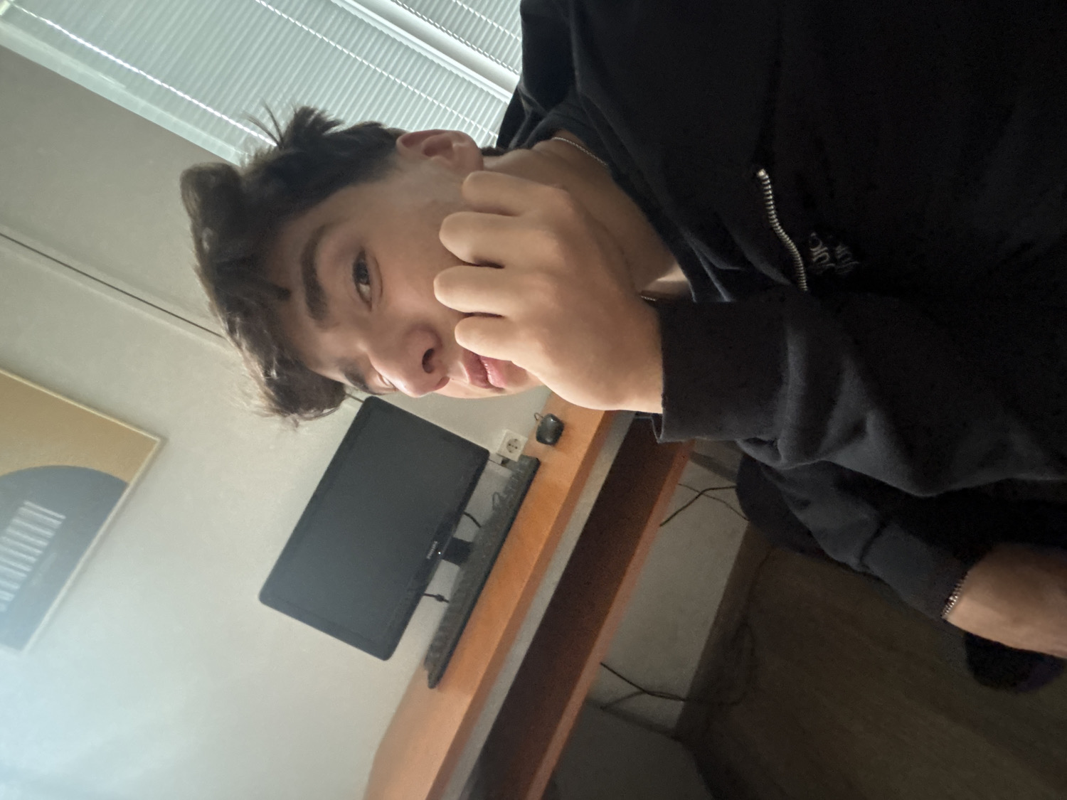 A young individual, appearing to be male, is featured in a close-up, rotated photograph taken indoors, likely in Troyan, Bulgaria. The person has short, messy dark brown hair and dark eyebrows, and looks directly at the camera with a neutral to slightly curious expression. Their right hand is partially visible, with fingers gently touching their face near the chin or cheek.
The individual is wearing a black hooded sweatshirt, partially unzipped to reveal a silver chain necklace. Faint, light-colored text is visible on the chest of the sweatshirt, near the zipper, though its specific content is not clearly discernible.
The background reveals a simple room setup, possibly a home office or study. A light-colored wooden desk is visible, on which rests a black computer monitor, a black keyboard, and a black mouse. A white wall socket is also present on the wall above the desk. To the left, a framed piece of abstract art hangs on a light-colored wall, depicting a dark semi-circle and horizontal lines on a tan background. Natural light filters into the room from a window with horizontal blinds, suggesting the photograph was taken during the daytime.