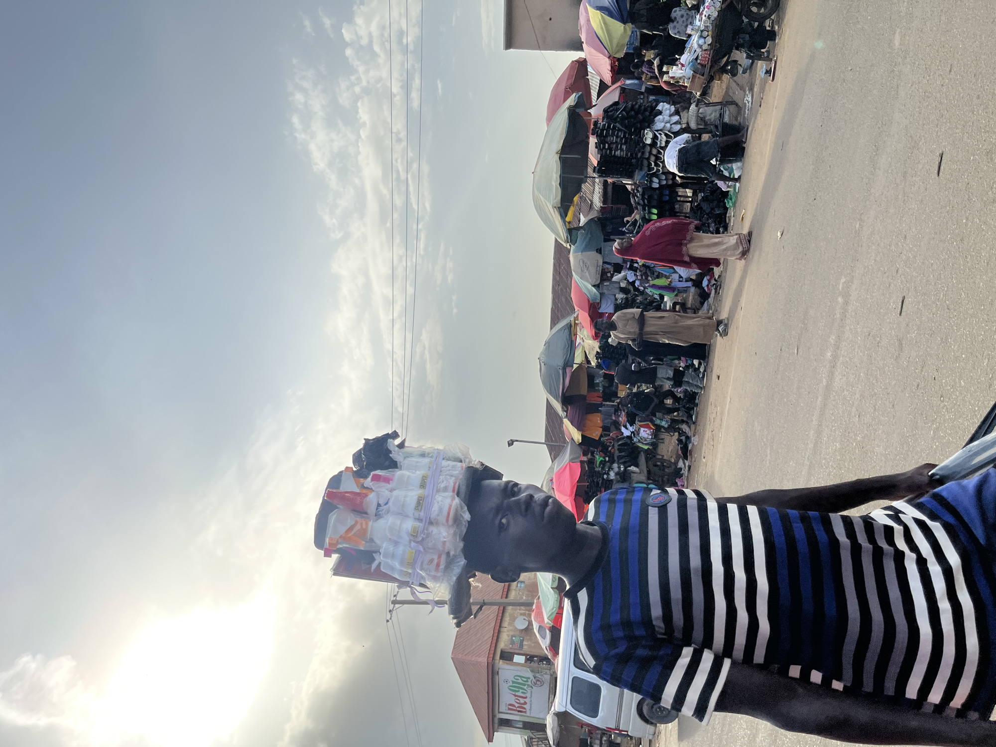 This media file shows a street scene in Rafin Par, Nigeria. The main subject is a young person, likely male, carrying a large load of goods balanced on their head. The load appears to consist of numerous small plastic containers, possibly filled with liquids or powders, wrapped in plastic. The person is wearing a horizontally striped t-shirt with black, white, and blue colors. They are walking on a paved road, and to the left of the frame, there is a street market with stalls covered by colorful umbrellas and awnings. Various goods are visible in the market stalls, including what appears to be footwear and clothing. Other people are present in the market, some browsing, and others tending to the stalls. In the background, there is a building with a red roof and a sign that reads "Bet9ja". The sky is partly cloudy, and the sun is visible, suggesting it is daytime, possibly late afternoon or early morning due to the angle of the light. The overall atmosphere appears busy and typical of a bustling marketplace.