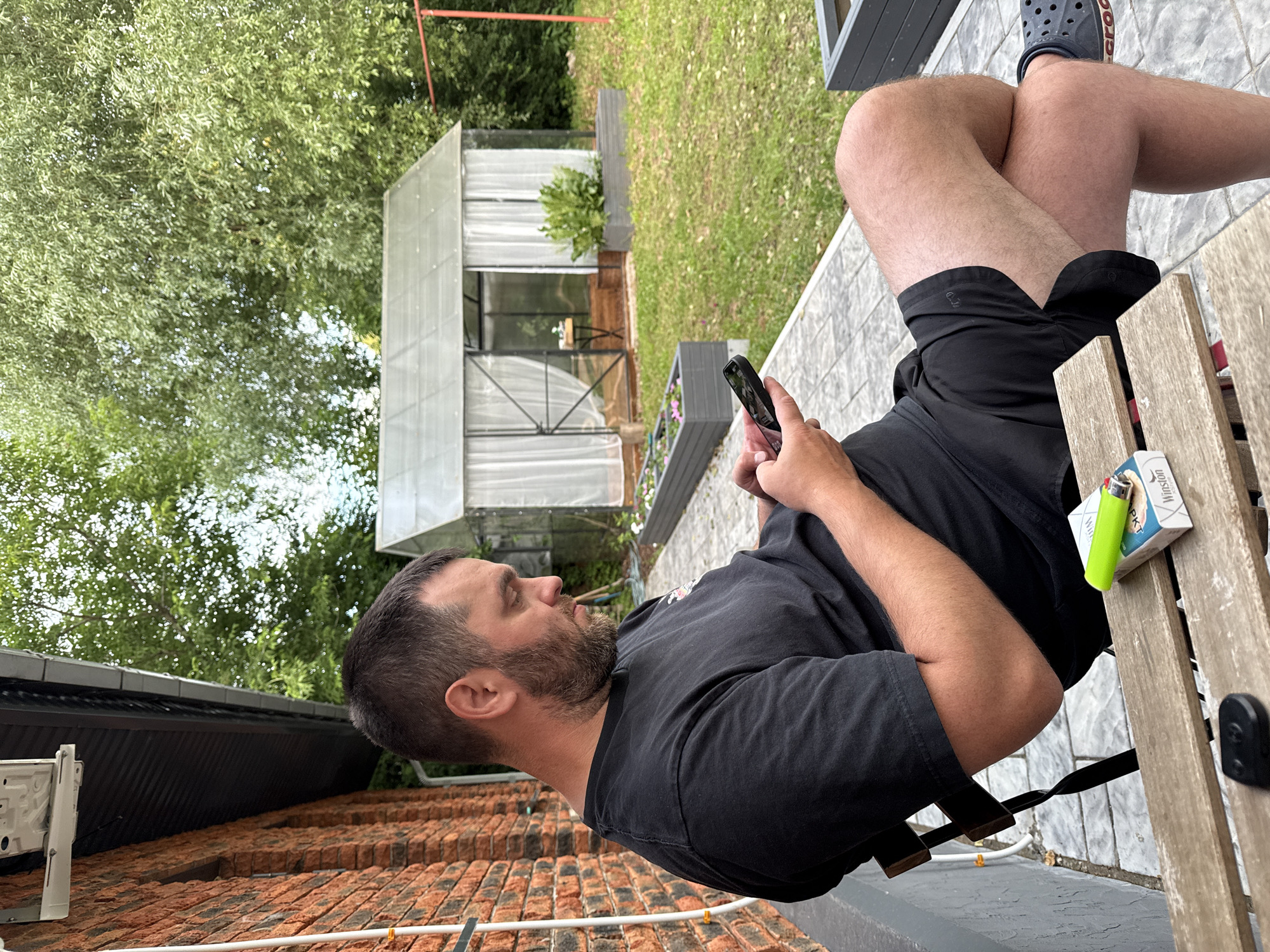 This media file shows a person sitting on a chair outdoors, holding a smartphone and a cigarette pack. The person has a beard and is wearing a black t-shirt and black shorts. A lighter sits on the wooden table next to the cigarette pack. In the background, there's a greenhouse, lush green trees, and a grassy area. The overall setting appears to be a backyard or patio area. The lighting suggests it is daytime. Visible text on the cigarette pack reads "Winston". The orientation of the image is rotated 90 degrees counter-clockwise.