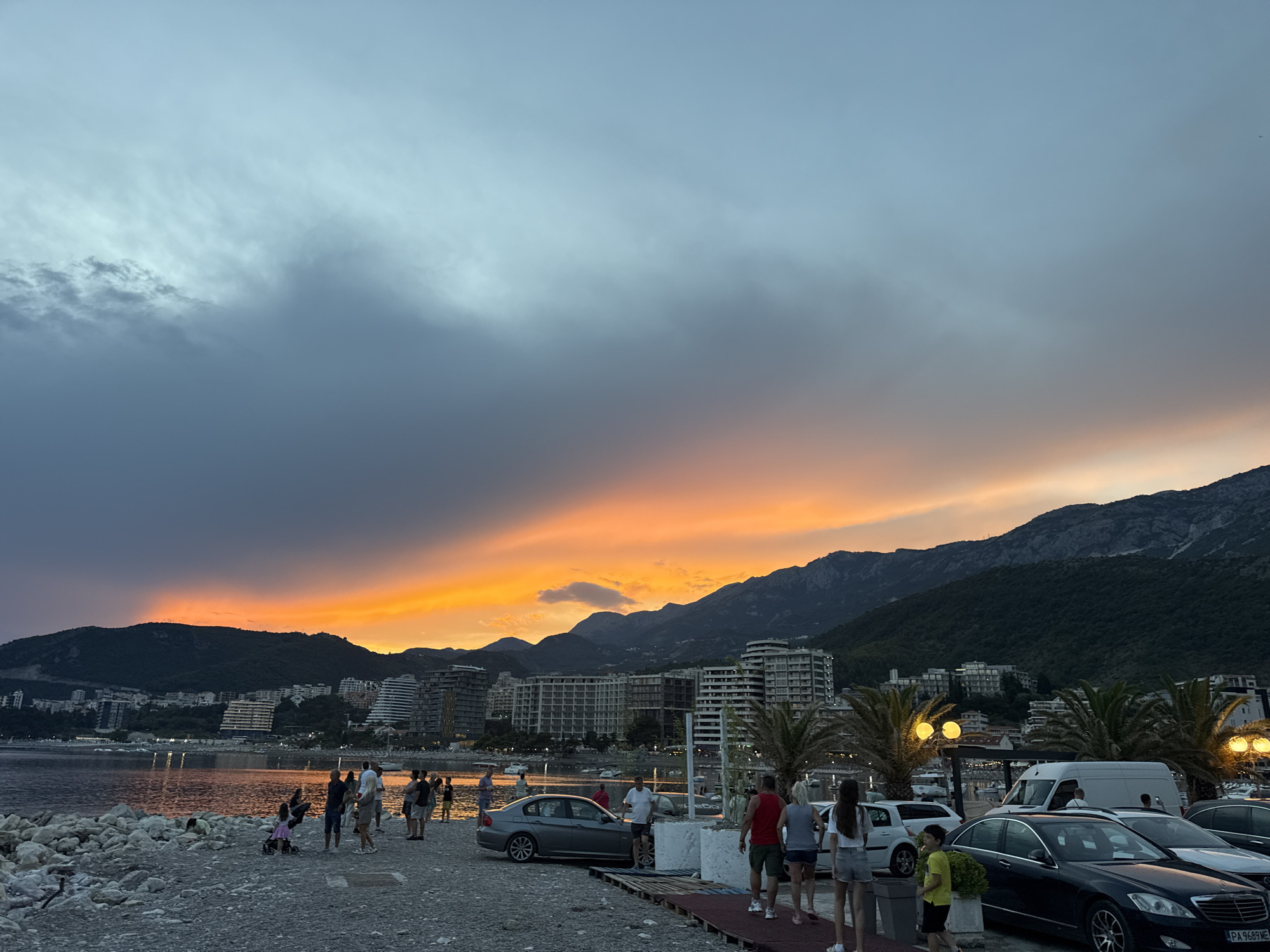 A scenic view of the coast of Bečići, Montenegro at dusk, with a vibrant sunset painting the sky with hues of orange, yellow, and grey. The calm sea reflects the colorful sky, while silhouetted mountains form a dramatic backdrop. Along the shore, a promenade is lined with people enjoying the evening. Modern buildings, some with unique architectural designs, are visible along the coastline, indicating a developed resort area. Palm trees and lampposts add to the atmosphere of a pleasant evening.
Several people are casually walking along the promenade, some appearing to be families or groups of friends. A few cars are parked nearby, including a black sedan and a white van. The overall scene suggests a relaxed and enjoyable atmosphere, perhaps a holiday or evening stroll by the sea. The time of day is clearly sunset or twilight, given the sky's colors and the lighting. The weather appears to be calm and clear, allowing for a beautiful sunset display. The presence of buildings and the mountainous terrain strongly suggest a coastal Mediterranean location, consistent with Bečići, Montenegro. The license plate of the black sedan is partially visible and appears to read "PA 9689 ME".