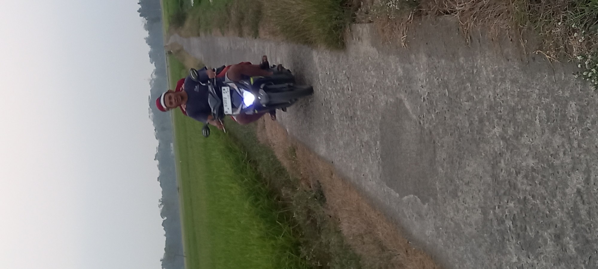 In Sidamulya, Indonesia, two individuals are riding a motorcycle on a paved path bordered by a lush green rice field and dry grass. The person in the front, wearing a dark t-shirt and a red and white hat, is operating the motorcycle. Behind them, another person, partially obscured and wearing a red garment and a similar hat, is seated as a passenger. The motorcycle's headlight is illuminated. The sky is overcast and light.