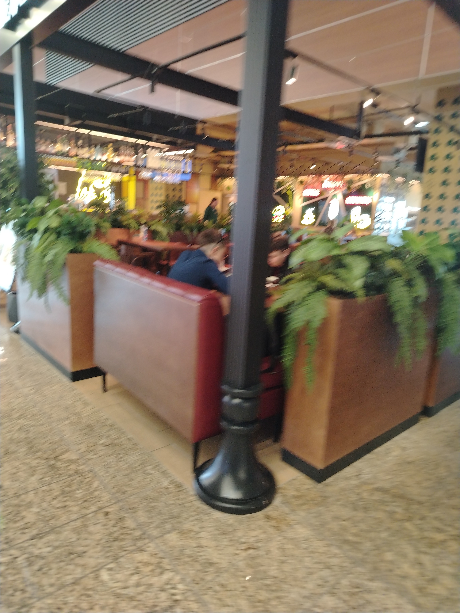 The interior of a restaurant in Khimki, Russia, features booth seating with brown faux leather upholstery. The booths are divided by planters filled with greenery. People are seated in the booths, and tables are visible in the background. The floor is a light-colored tile. A dark grey support column is in the foreground. Overhead, there is a combination of black metal beams and a slatted ceiling. Various signs and lighting fixtures are visible in the background.