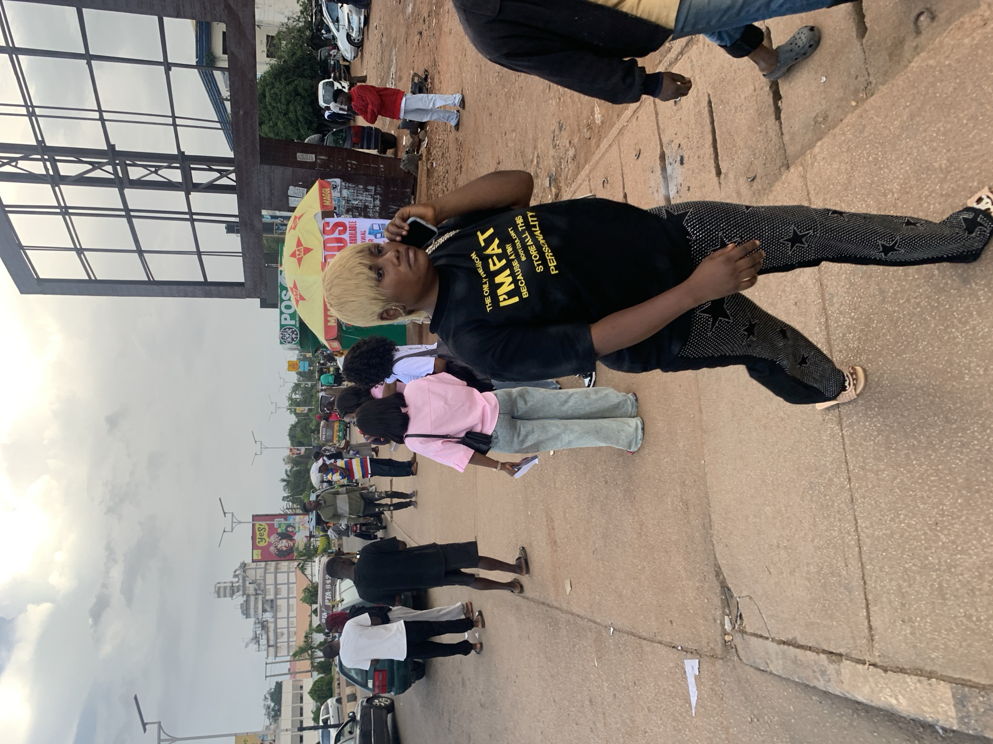 This media file shows a street scene in Jos, Nigeria, likely during the daytime given the overcast sky. The foreground features a person with blonde, spiked hair, wearing a black t-shirt with yellow text and black pants adorned with white stars. They are holding a phone to their ear and appear to be engaged in a conversation. Behind them, a group of people are gathered, some wearing colorful clothing. In the background, there are buildings, billboards, and various vehicles, including cars and motorcycles, suggesting a busy urban environment. One billboard displays the text "Yes!" and other promotional content related to "POS," "MAGGI," and "AVAILABLE." The overall atmosphere seems casual, with people walking and interacting on a paved street. The weather appears cloudy, indicating a possible rainy or overcast day.