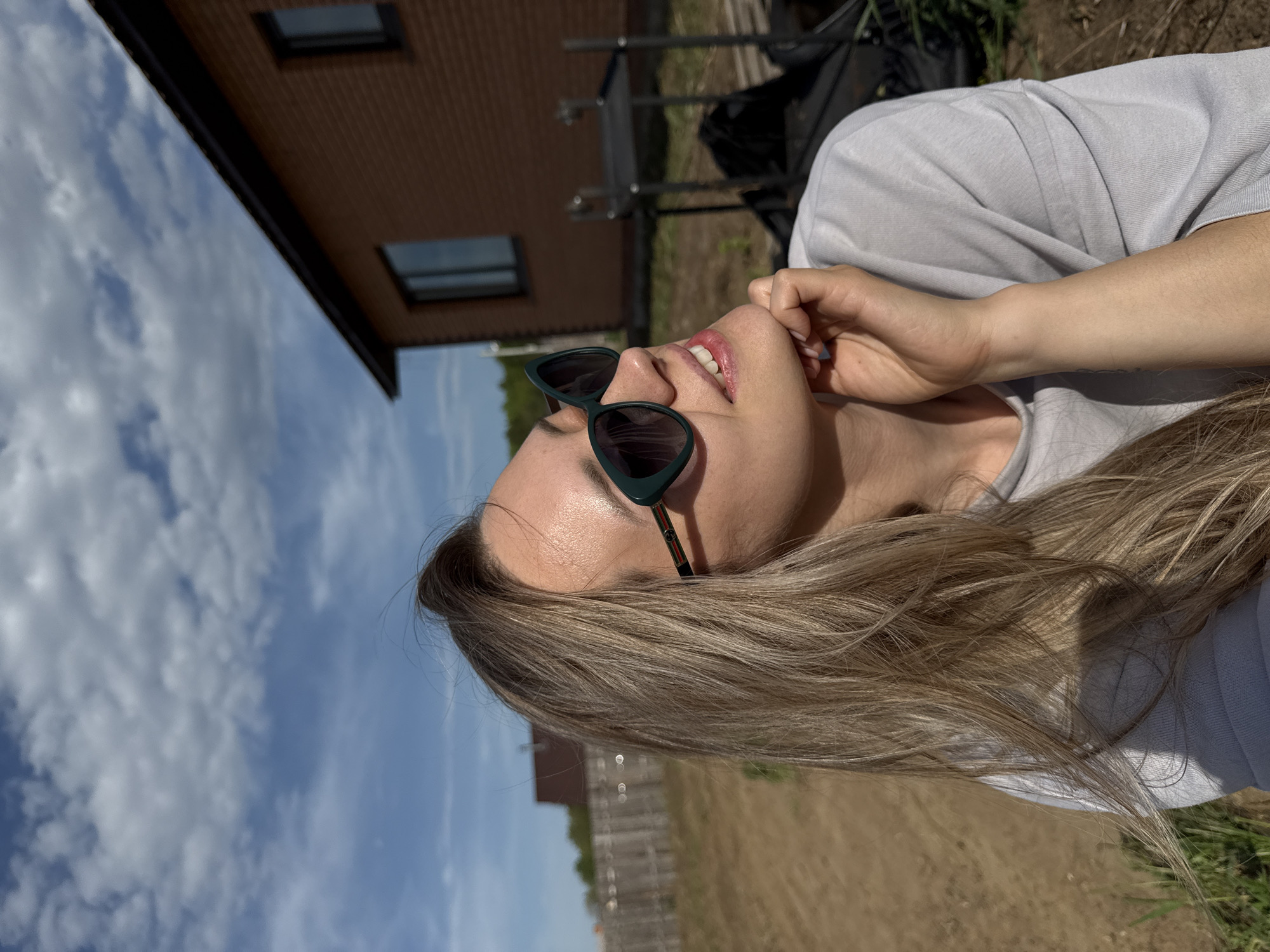 A woman is outdoors wearing dark sunglasses and a light grey shirt. She is looking up and to the side with her right hand near her face. Behind her, there is a building with visible windows. The sky is blue with clouds. There is also a grassy and dirt area, possibly a yard, behind the woman. The specific location in Chaikovsky, Russia, cannot be determined from the provided image.