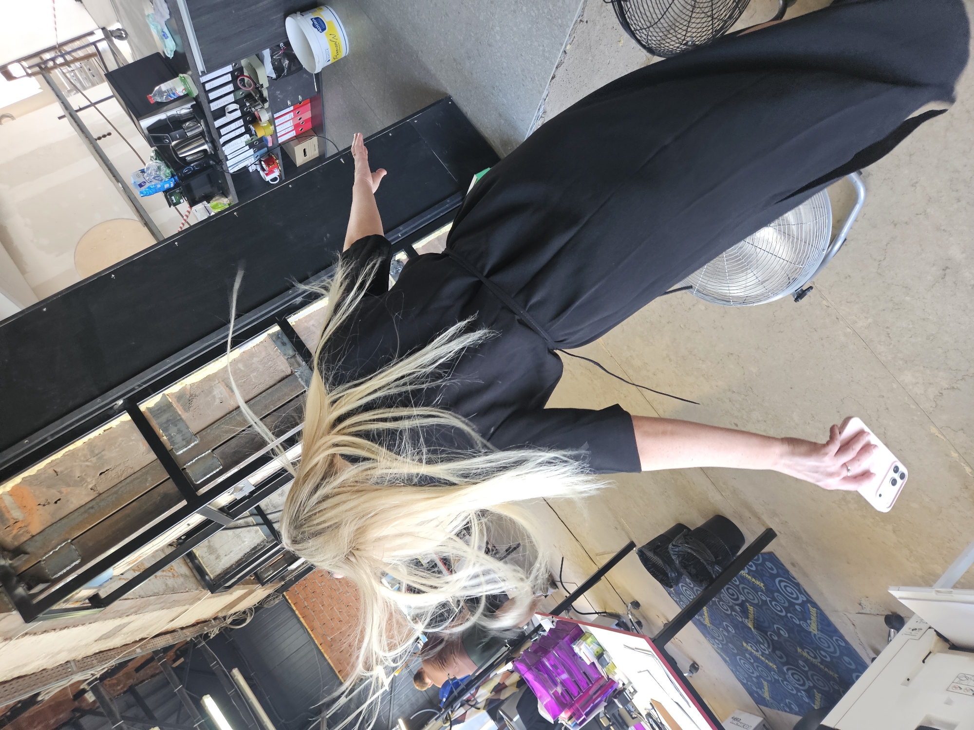 A woman with long, blonde hair is standing in what appears to be an indoor location. She is wearing a black dress with short sleeves and a thin tie around the waist. Her right arm is extended, holding a mobile phone. Her left arm is also extended upwards. In the background, there are shelves with various items, including containers, binders, and equipment. The environment has a mixed aesthetic, with some areas looking unfinished and others appearing more organized. There are also fans visible in the background. The overall location cannot be identified as being in Chaikovsky, Russia from the visual information provided.