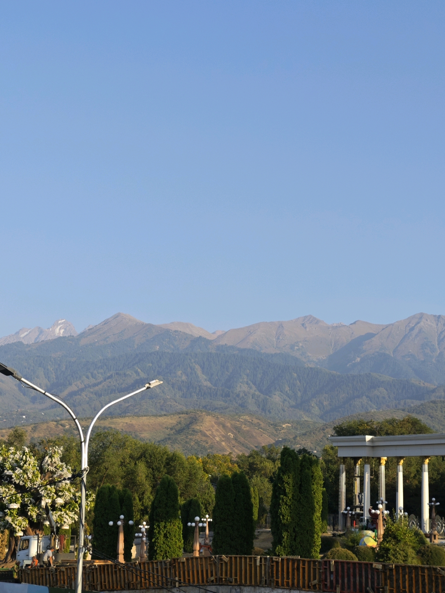 The image shows a scenic view of the mountains in Almaty, Kazakhstan. In the foreground, there are streetlights, lush green trees, and a white structure with columns that appears to be a part of a park or an architectural monument. A white truck and a few people are visible near the truck, suggesting ongoing activity or maintenance work. The background is dominated by the majestic Tien Shan mountain range, with forested slopes leading up to rocky peaks, some of which are snow-capped. The sky is clear and blue, indicating a pleasant day. The overall scene evokes a sense of natural beauty and urban development coexisting.