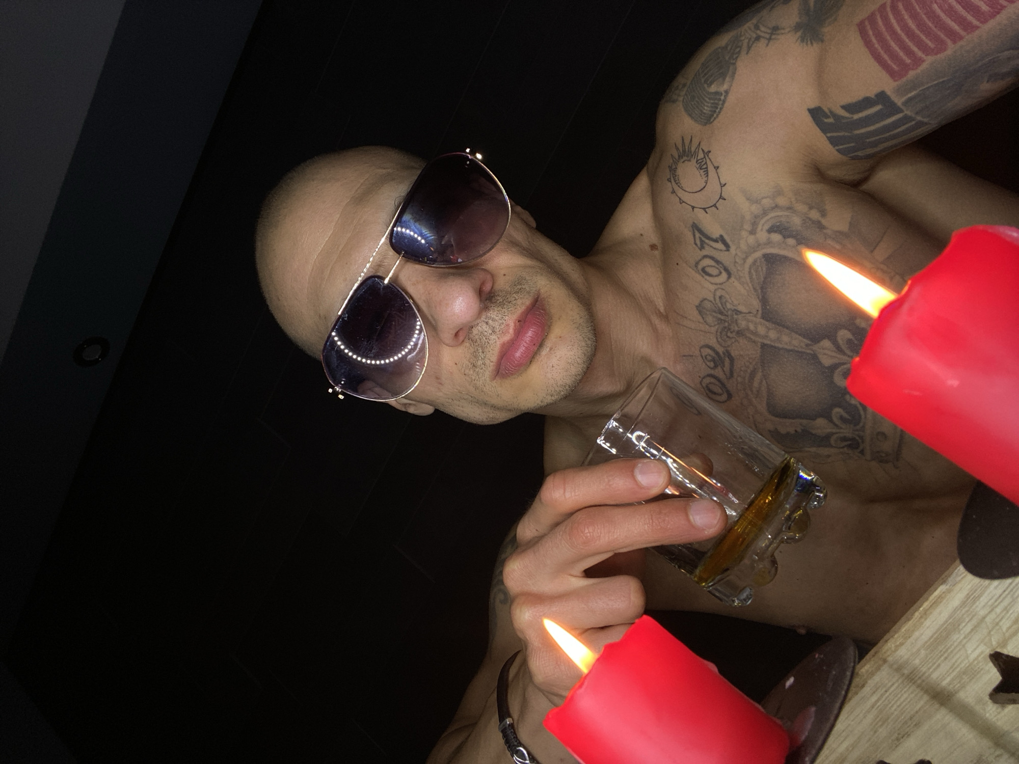 A low-angle shot shows a bare-chested man with tattoos wearing aviator sunglasses. He holds a glass of amber liquid. Two lit red candles flank him, illuminating a wooden surface. The background is dark. It is not possible to determine if the photo was taken in the city of Pleven, Bulgaria from visual information.