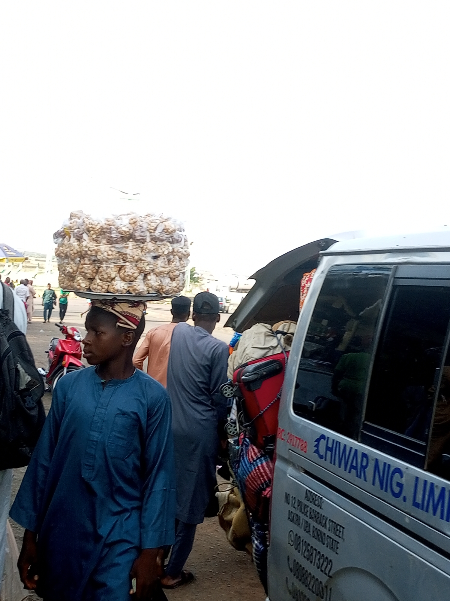 This is a medium shot, eye-level, outdoor photograph taken in the city of Gombe, Nigeria. In the foreground, a young boy wearing a blue robe and a patterned headwrap stands facing left, with a large tray of bagged goods balanced on his head. Behind him, other people are visible, some wearing traditional clothing. A red motorcycle is parked to the left of the boy. To the right, a silver van is partially visible with its rear door open, revealing luggage inside. Text on the van reads "CHIWAR NIG. LIMI" and includes an address "NO 12, POLICE BARRACK STREET, ASKIRA/UBA, BORNO STATE" and phone numbers. The ground appears to be dirt or gravel, suggesting a market or street setting. The overall atmosphere is bustling and appears to be daytime due to the bright sky.