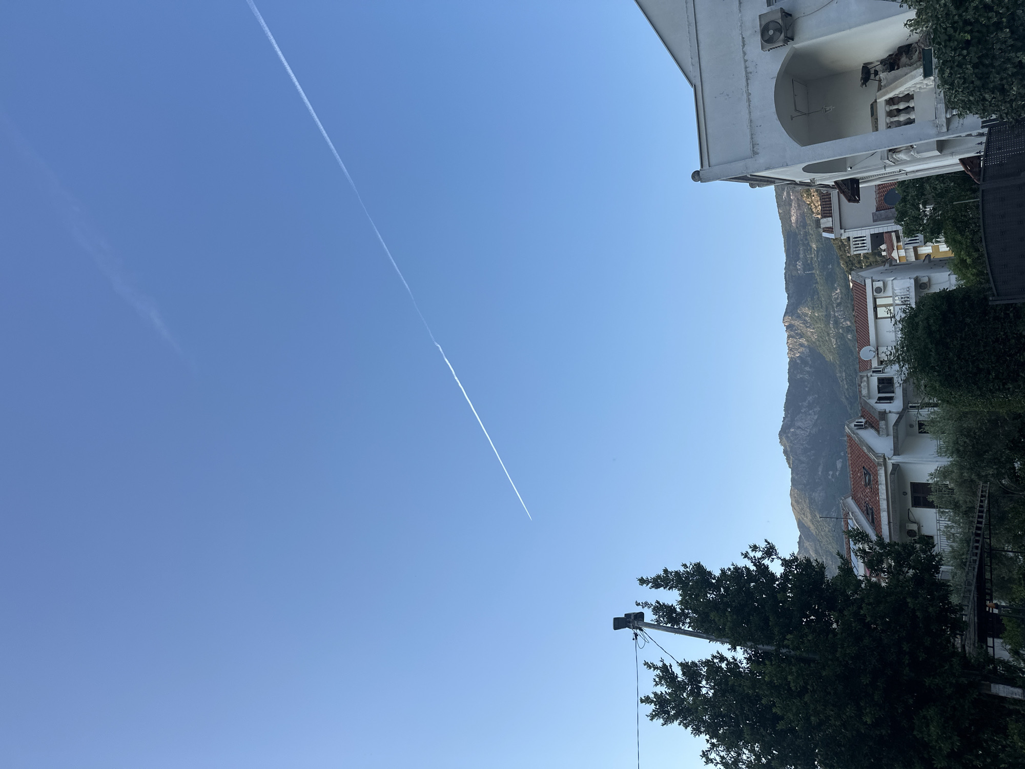 The image shows a view in Budva, Montenegro. The sky is clear blue with two contrails visible. Buildings with white facades and red tile roofs are present, partially obscured by trees. In the background, a mountain rises. A utility pole and wires are also visible near a tree.