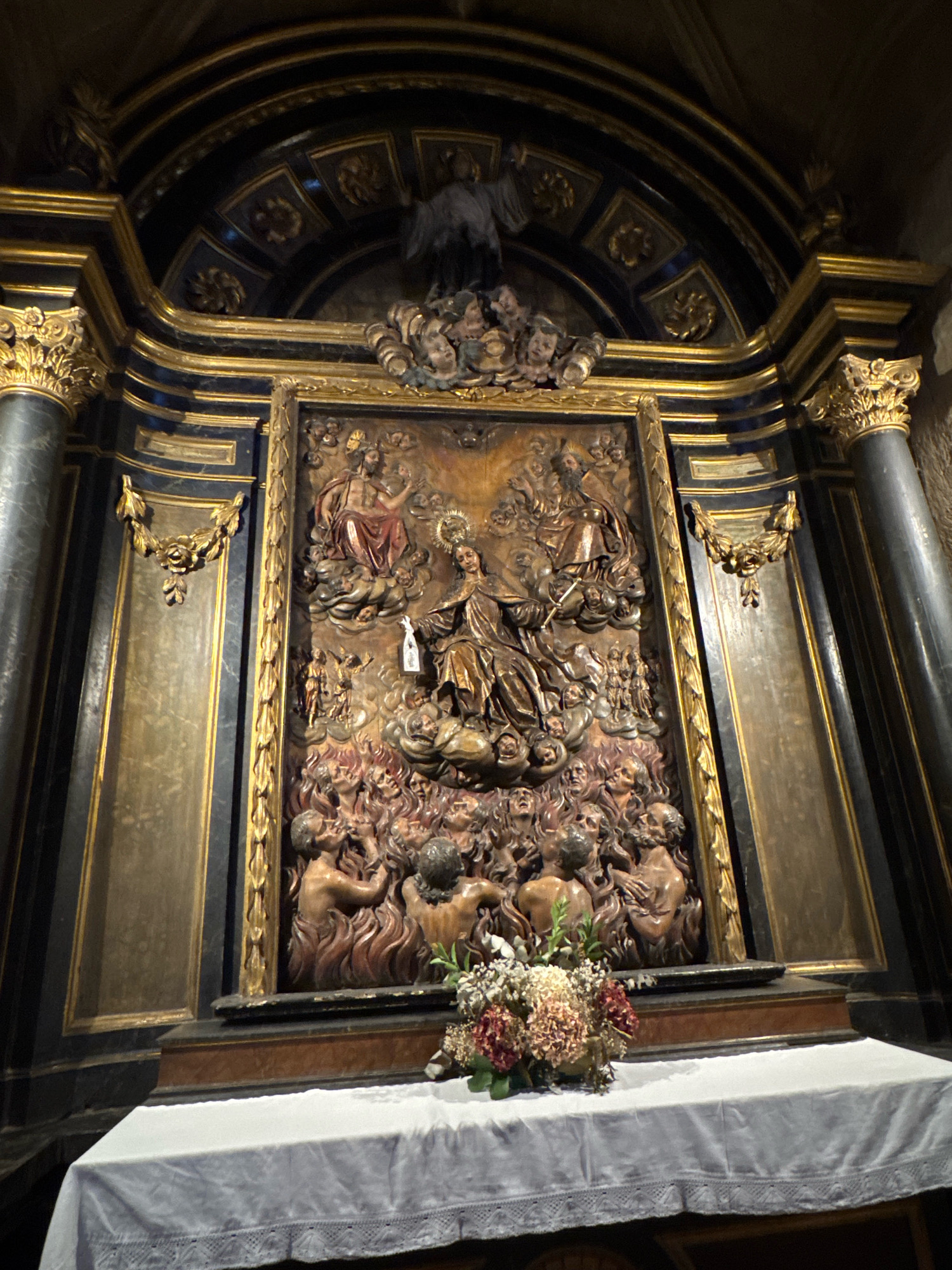 This image depicts an ornate religious altar with a large, carved wooden panel as its centerpiece. The panel shows a complex scene of figures, possibly representing a religious narrative. At the top, there are figures seated among clouds, with one figure in the center holding a staff and appearing to be in a commanding position. Below this, there are smaller figures, some appearing to be in prayer or supplication. The lower portion of the panel depicts a scene of souls in flames, with many contorted faces expressing torment or anguish.
The altar itself is elaborately decorated with gilded architectural elements, including columns, cornices, and decorative motifs. The dark background of the altar contrasts with the warm tones of the carved panel and the gold accents. In front of the altar, a white cloth-covered table holds a bouquet of dried flowers.
This altar is located within the San Vicente Church (Iglesia de San Vicente Mártir) in San Sebastián, Spain. The church is a prominent example of Gothic architecture in the city.