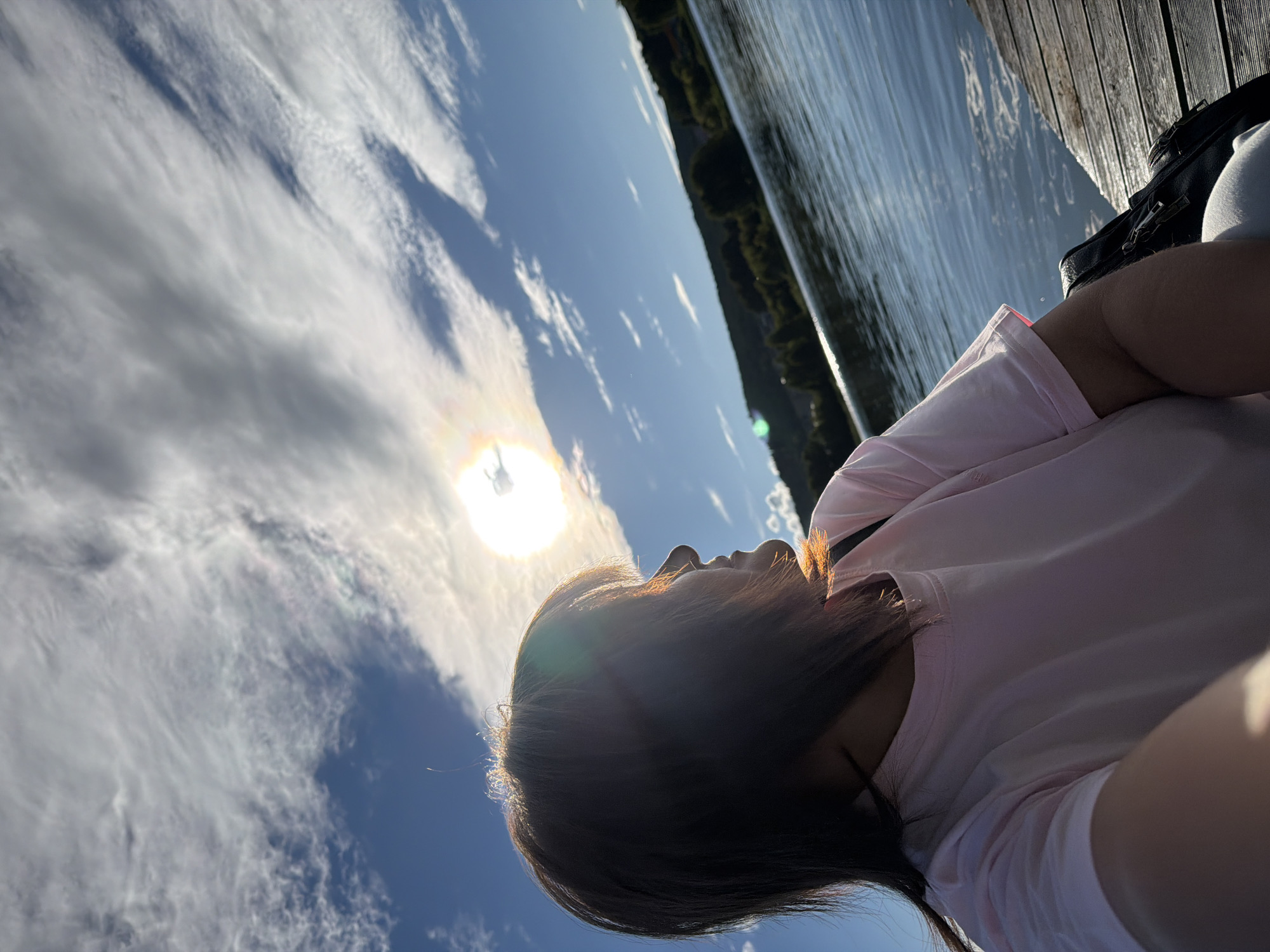 The image shows a person lying on a wooden deck next to a body of water, possibly a lake or river. The person is wearing a light pink t-shirt and their head is tilted back, looking up at the sky. Their hair is dark and frames their face, and the sun is shining brightly, creating a lens flare effect. The sky is blue with wispy white clouds, and the sun is partially obscured by clouds. The water is rippling, reflecting the sky and the surrounding trees which line the opposite bank. The wooden planks of the deck are visible in the foreground, with a dark bag placed on them. The overall impression is one of relaxation and enjoyment of a sunny day outdoors. The angle of the sun suggests it could be late afternoon or early morning. There is no visible text in the image.